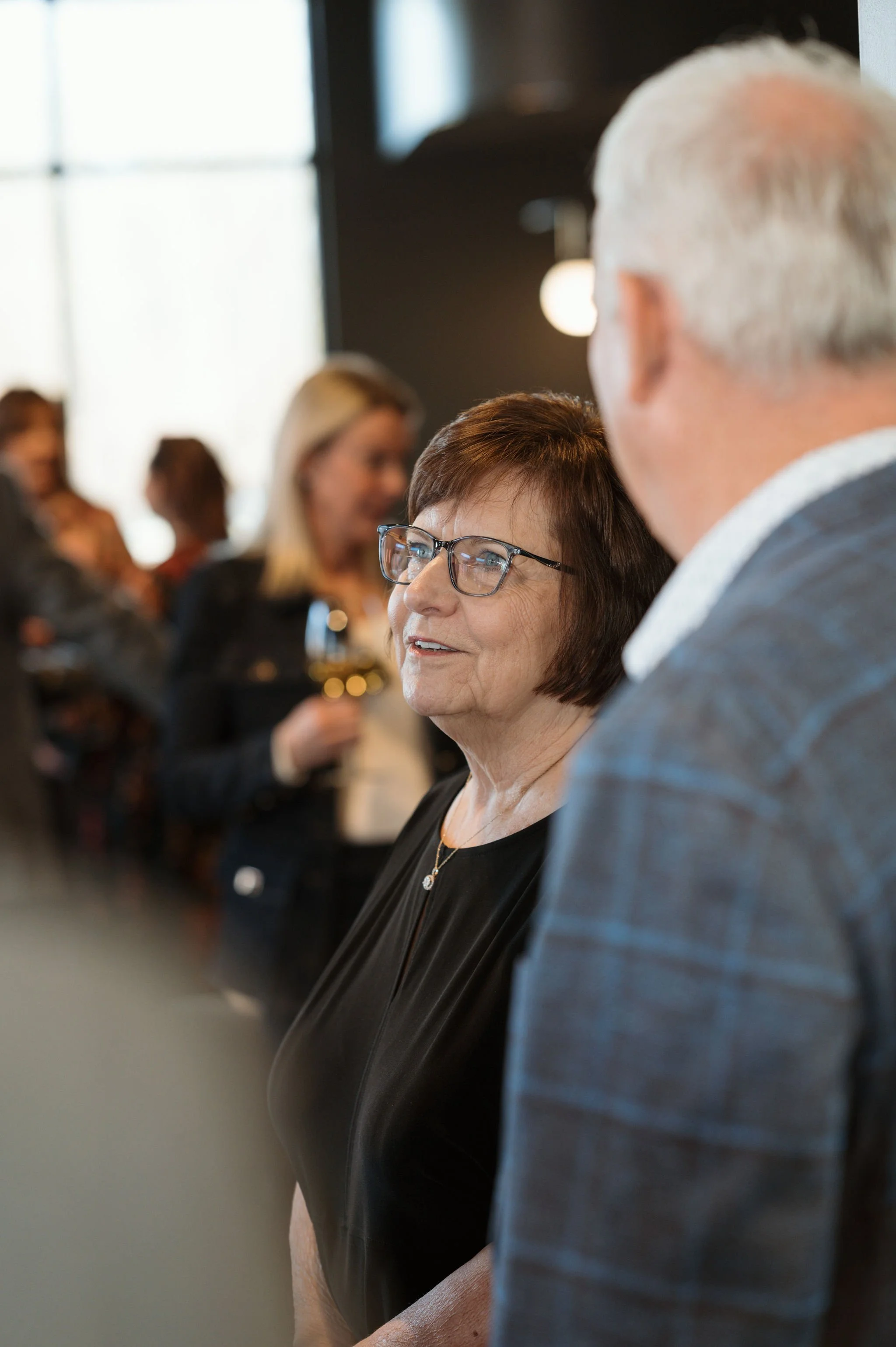 People socializing at a gathering or event, with a focus on a woman wearing glasses and a man in a suit engaging in conversation, blurred background with other attendees.