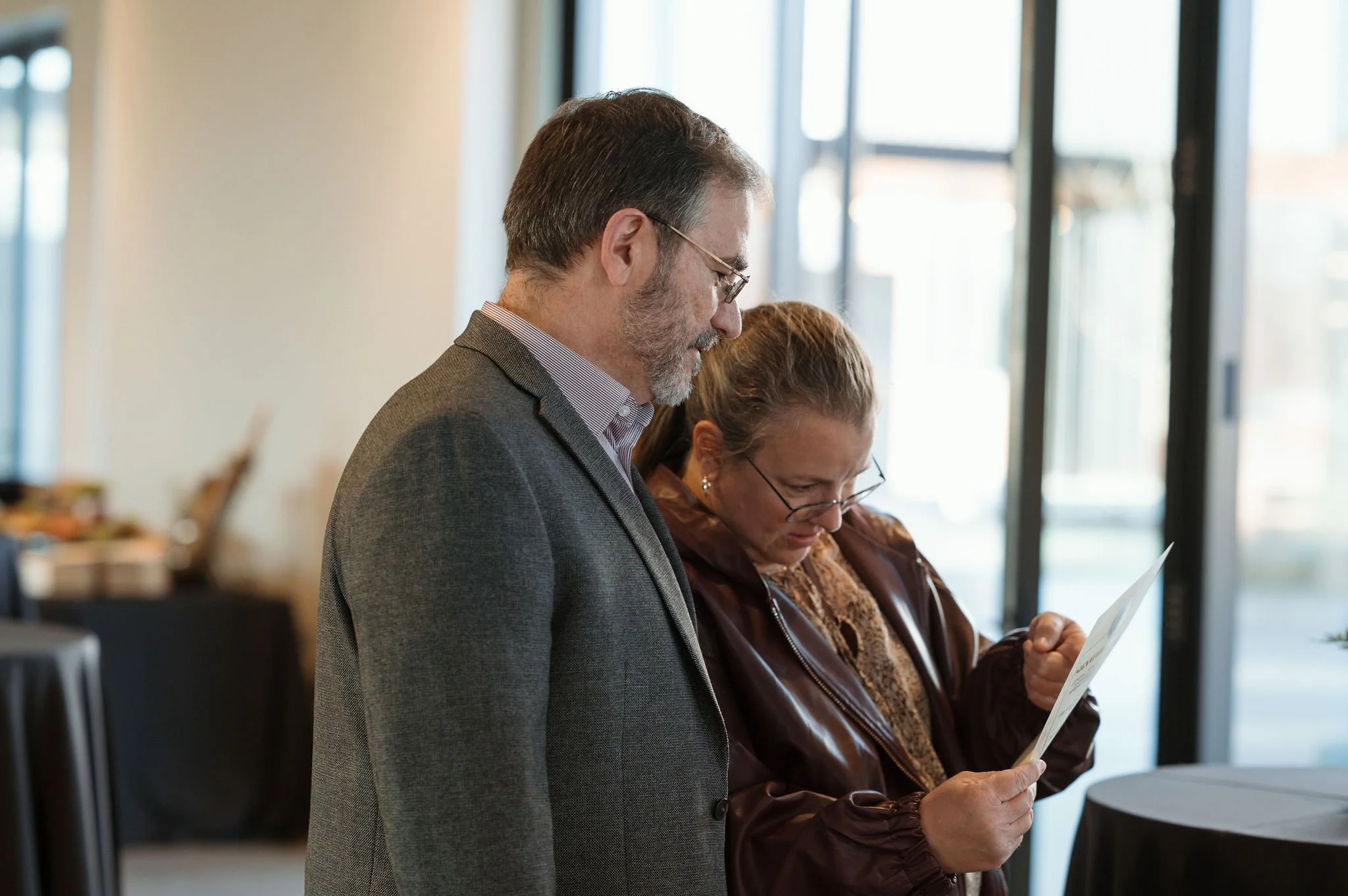 A man with glasses and gray hair wearing a gray blazer stands next to a woman with glasses and brown hair wearing a brown jacket, both looking at papers in an office with large windows.