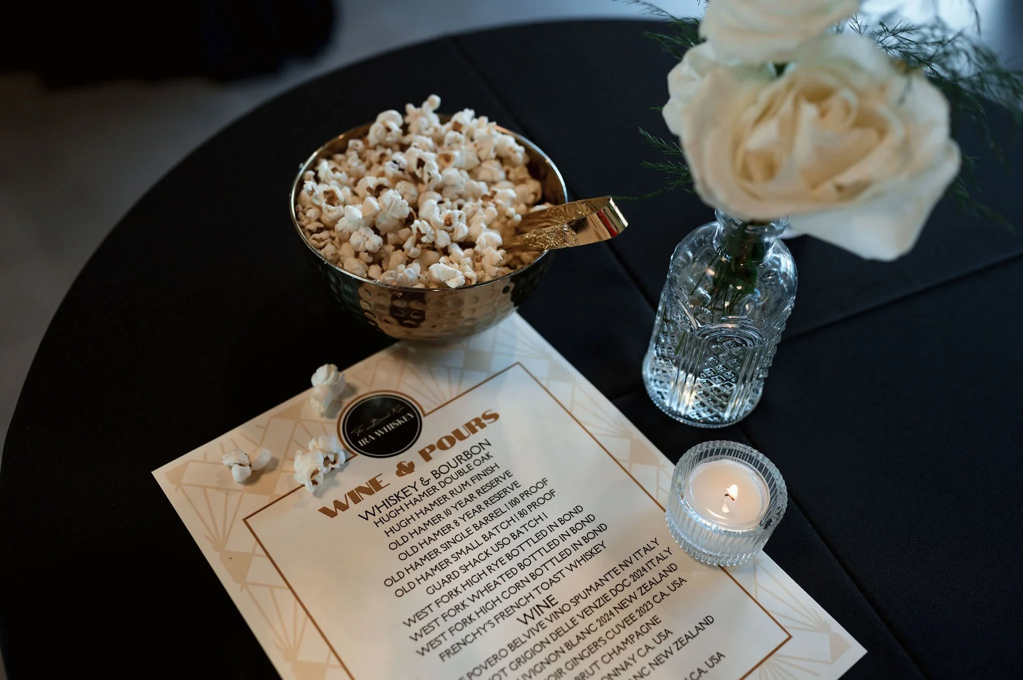 A table with a gold and black bowl of popcorn, a menu, a small lit candle in a clear glass holder, and a vase with white roses and greenery against a dark background.
