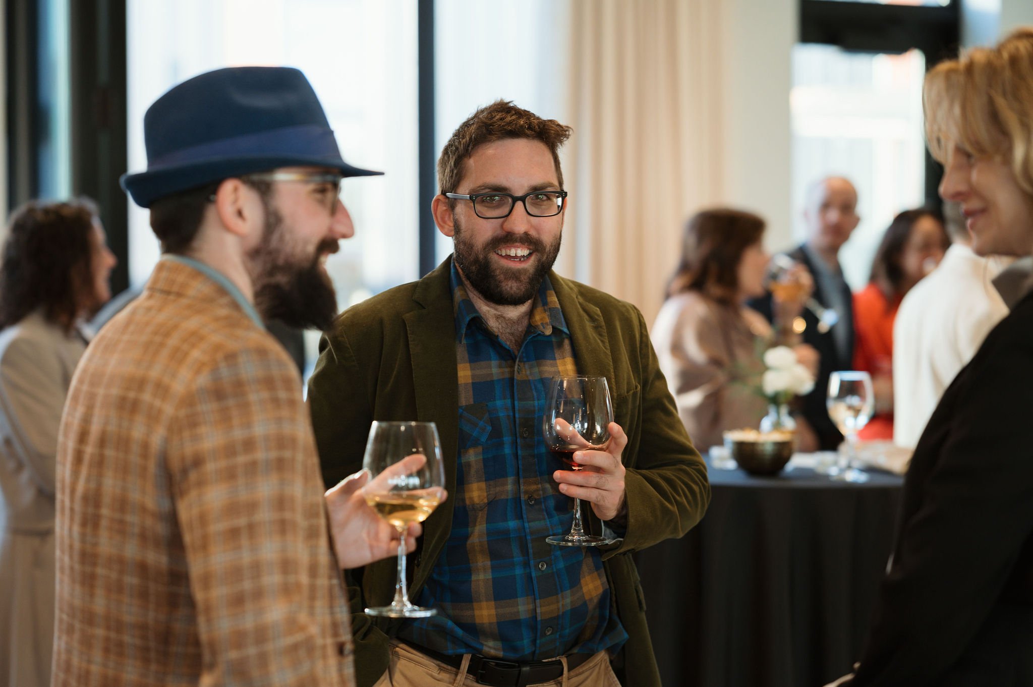 Group of people in a social gathering, with focus on a man with glasses and a beard holding a glass of white wine, talking to others in a well-lit room.