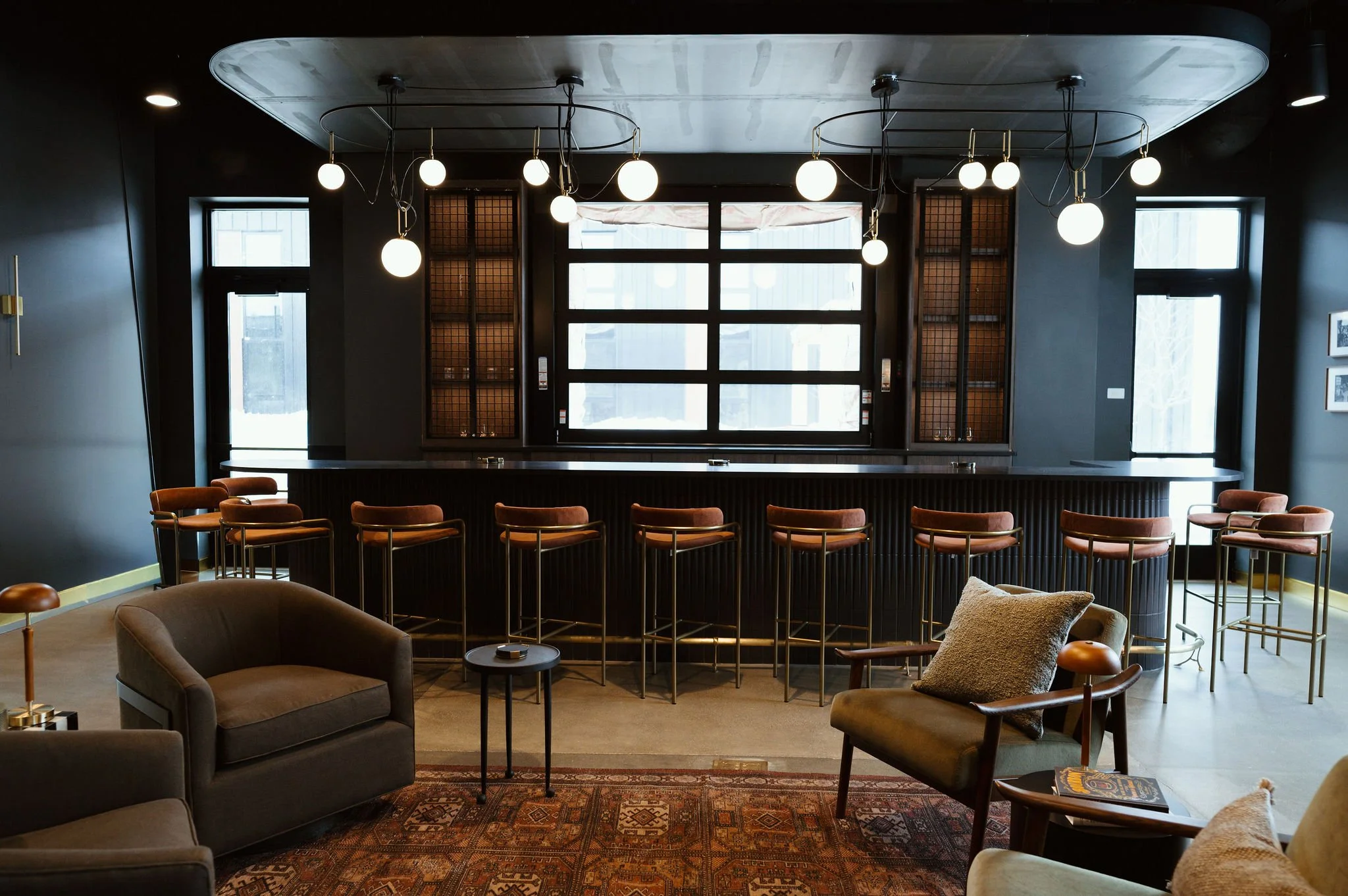 Interior of a modern lounge or restaurant with a dark color scheme, featuring large windows, hanging globe light fixtures, a bar counter with seating, and comfortable chairs and sofas.