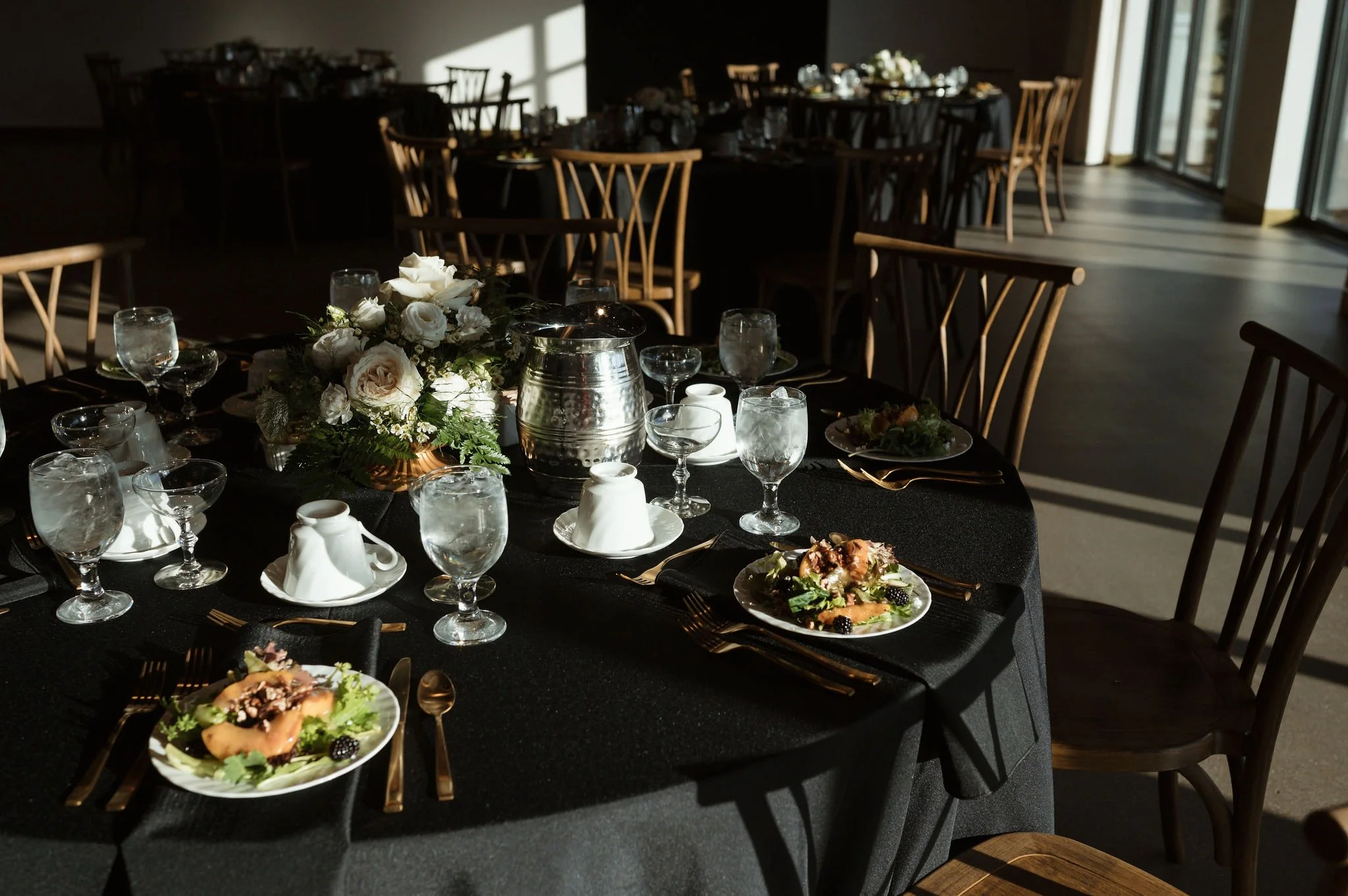 A dining table set with white flowers, glassware, and plates, in a room with sunlight streaming through glass doors.
