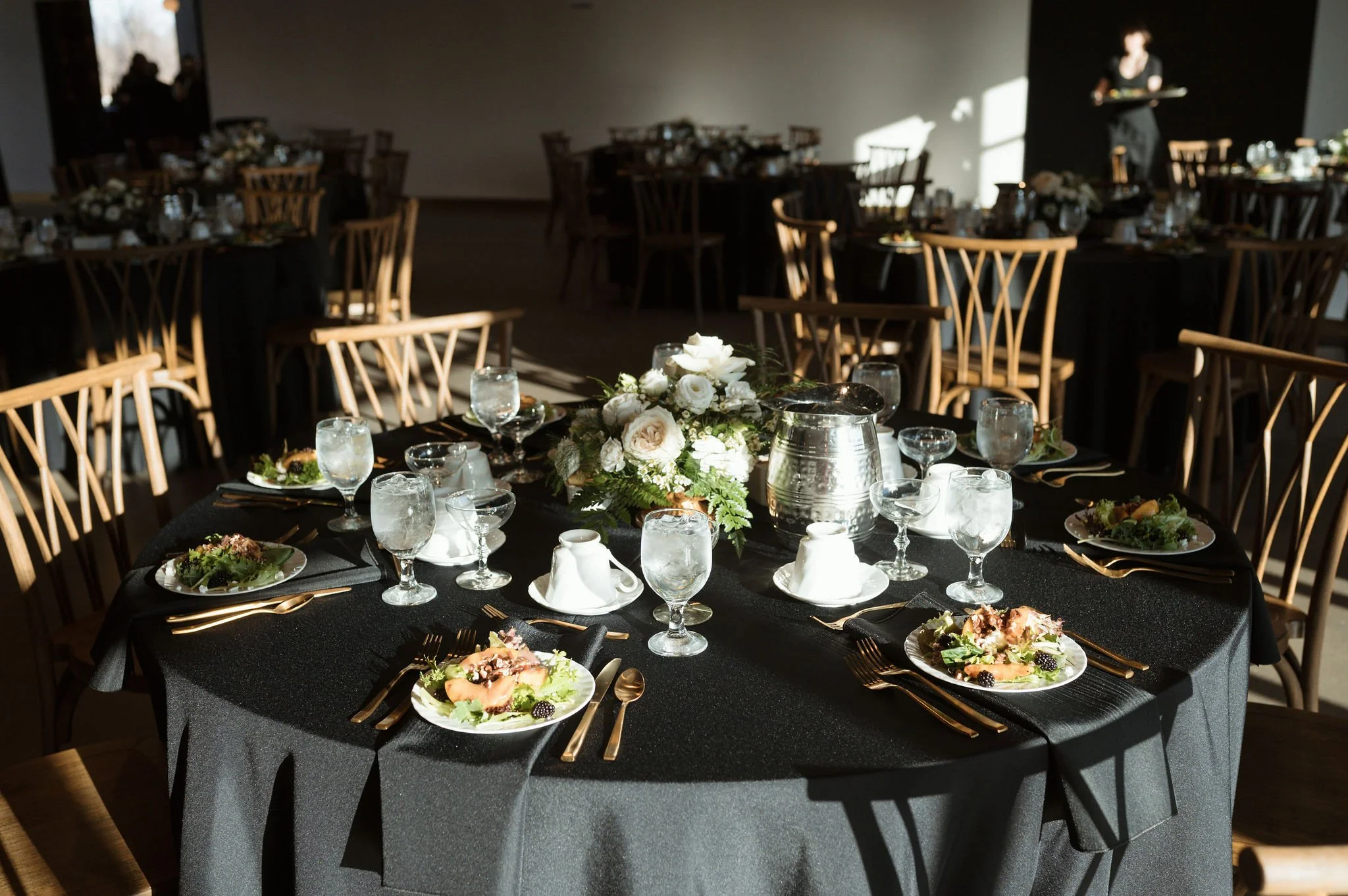 A banquet table set for a formal event with black tablecloths, floral centerpieces, and place settings including plates, glasses, and gold flatware. Chairs with wooden backs surround the table, and sunlight creates shadows on the wall.
