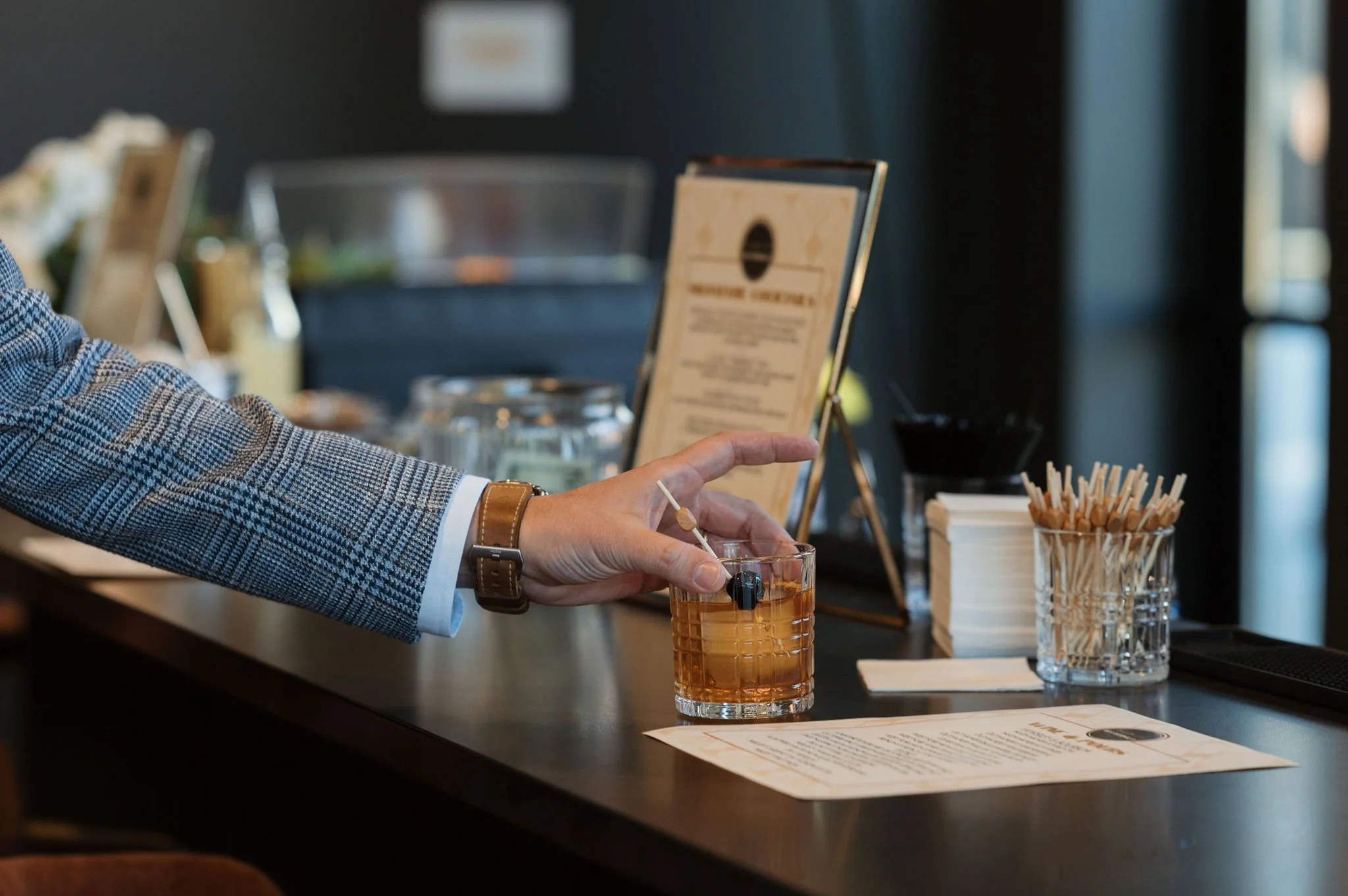 A person in a checkered blazer and brown watch is holding a beverage glass with an orange-colored drink and a skewer of cheese in a restaurant or cafe setting. The table has a menu, napkins, and a container of toothpicks, with blurry background detai