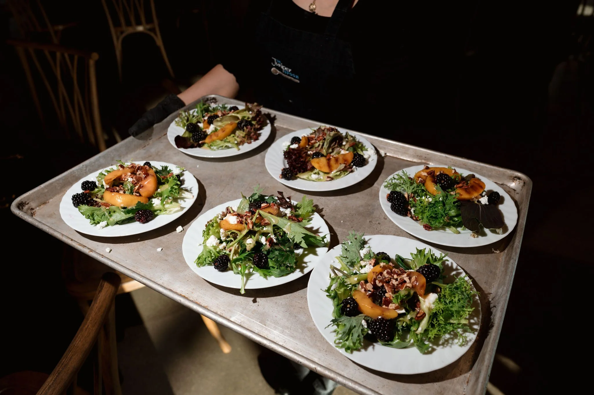 Multiple plates of salads with mixed greens, blackberries, sliced peaches, cheese, and nuts on a tray.