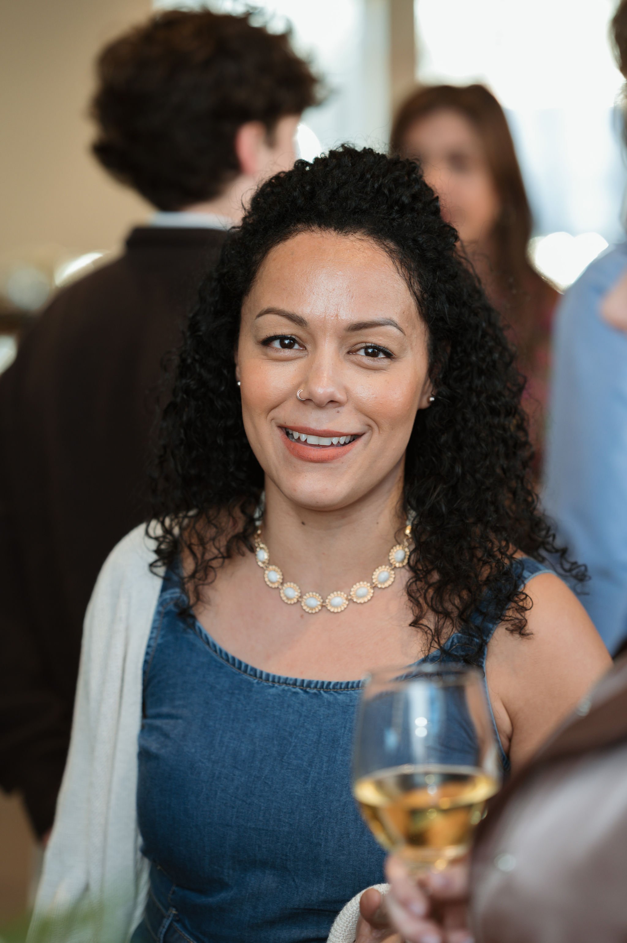 A woman with curly black hair and a nose piercing smiling at a social gathering, wearing a blue top with a white cardigan and a pearl necklace, holding a glass of white wine.