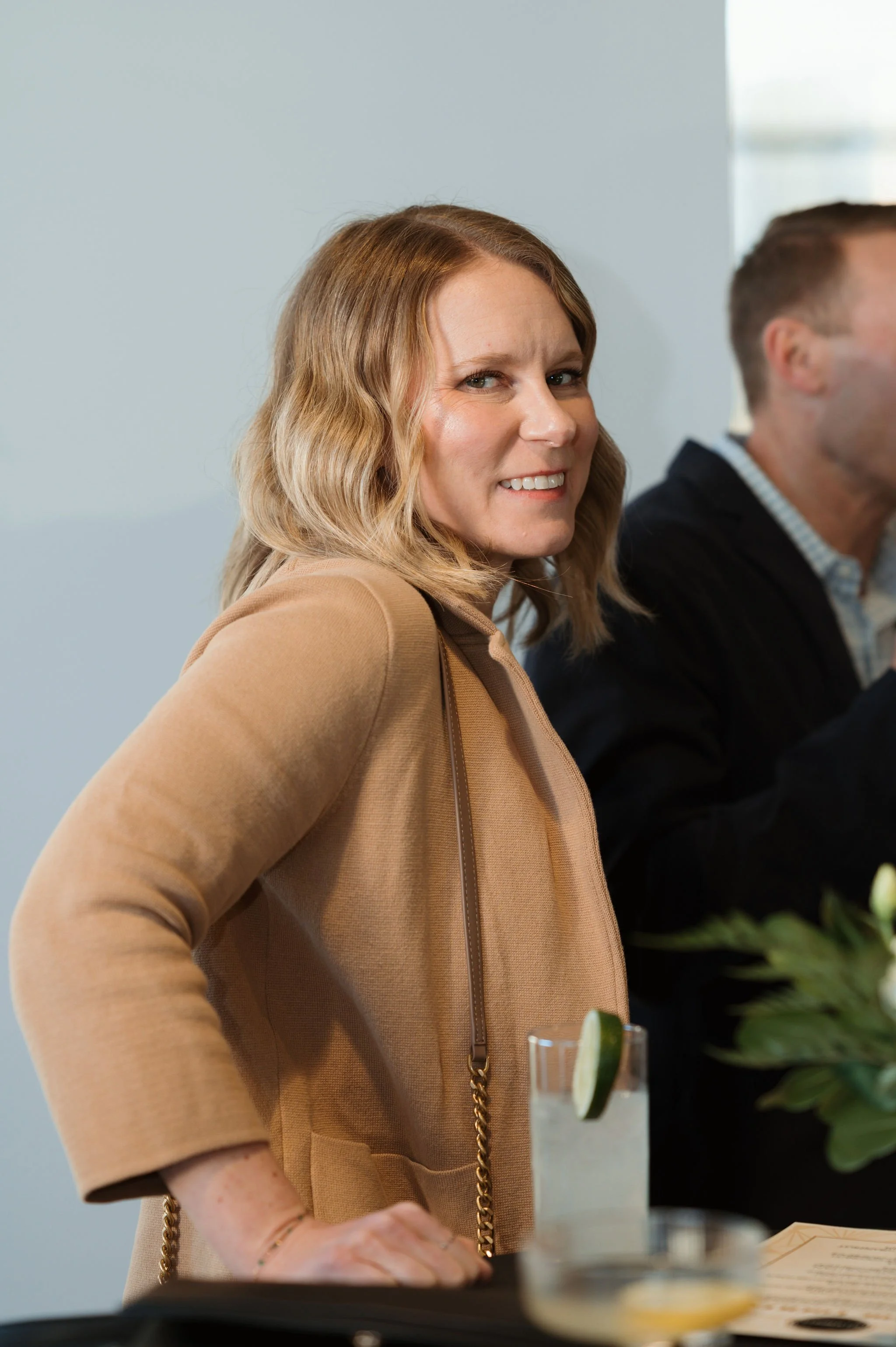 A woman with wavy blonde hair, wearing a tan jacket, smiling slightly, standing at a table with a drink garnished with lime slices and a menu or paper in front of her.