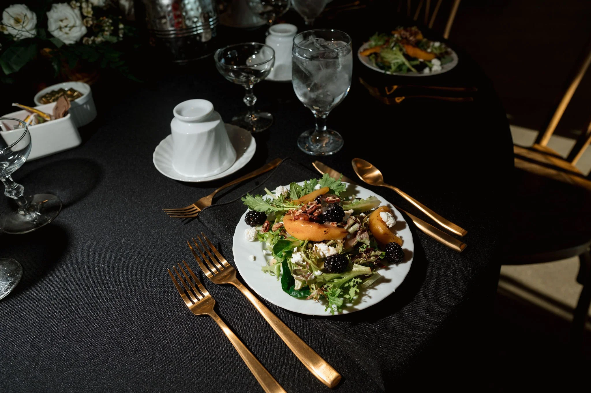 A dining table set for a meal with a black tablecloth, featuring a white plate of salad with greens, blackberries, feta cheese, and pear slices, gold utensils, and glasses of water, with floral decorations and additional plates and glasses in the bac
