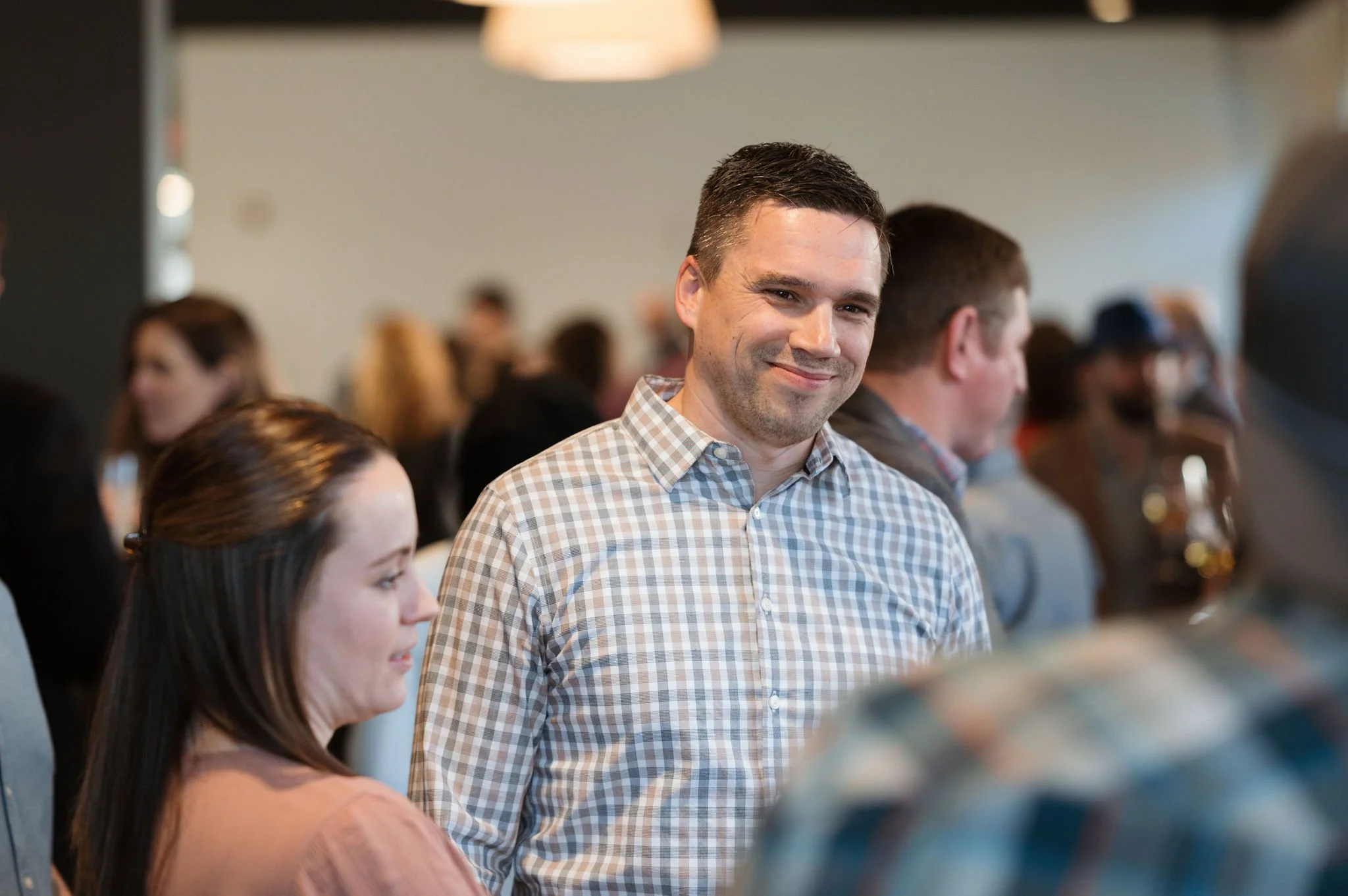 A man in a checkered shirt smiling at a social gathering with other people in the background.