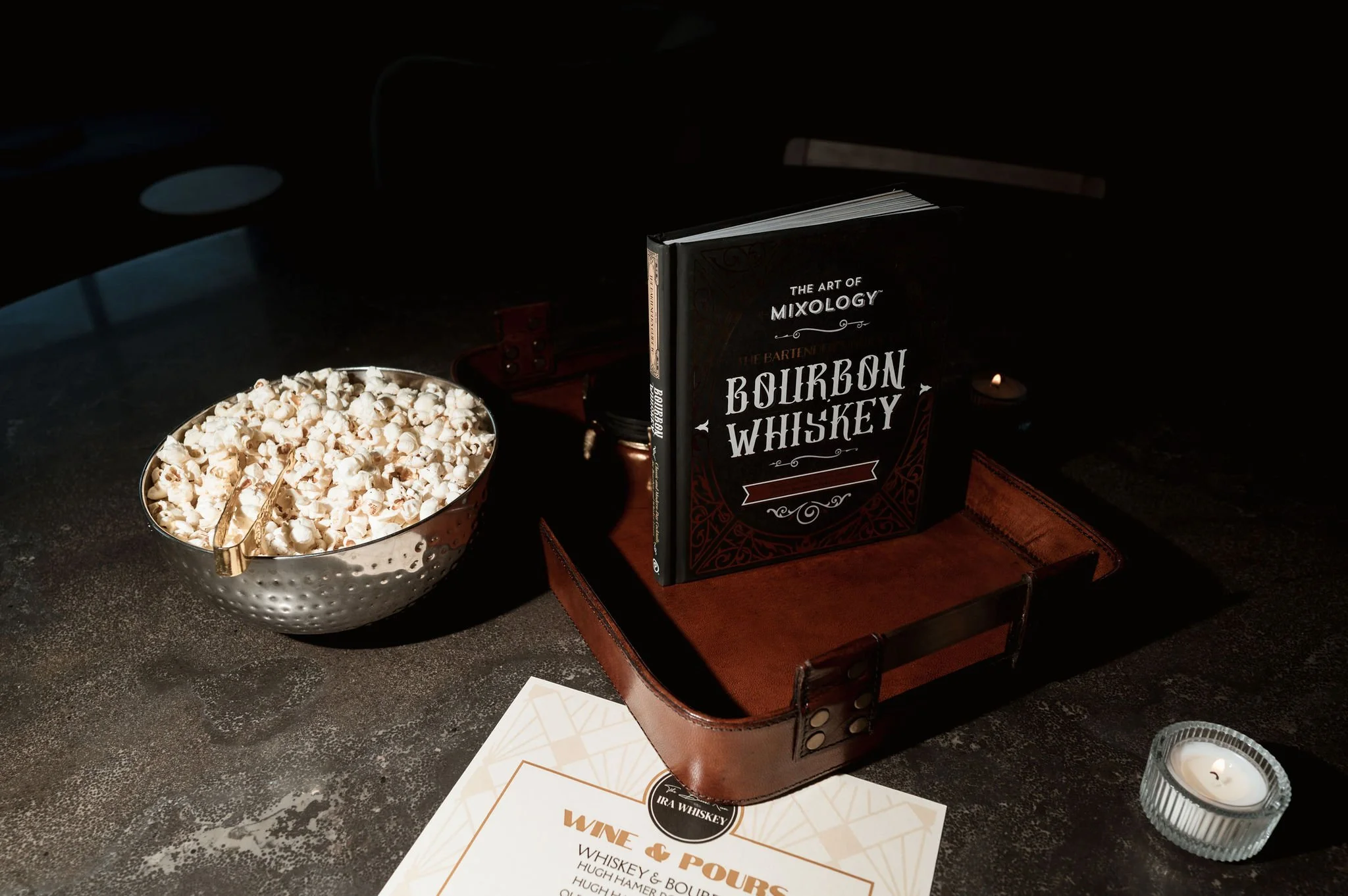 A book titled 'The Art of Mixology' with the subtitle 'The Bartender Bourbon Whiskey' is placed on a wooden tray. Next to it is a bowl of popcorn with a small pair of tongs, placed on a dark surface. There is also a candle in a glass holder below the