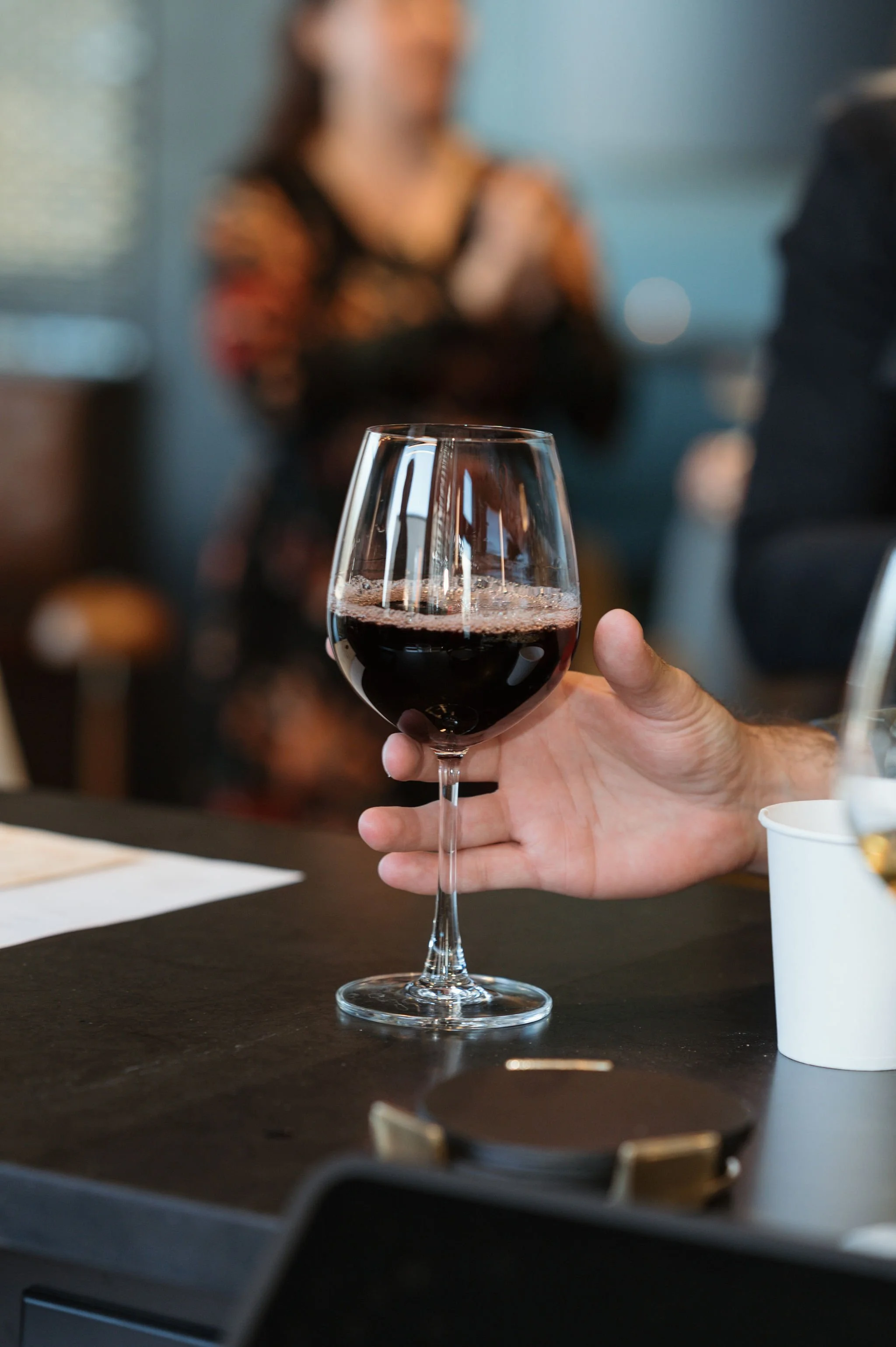 Person holding a glass of red wine in focus, with blurred woman in background.