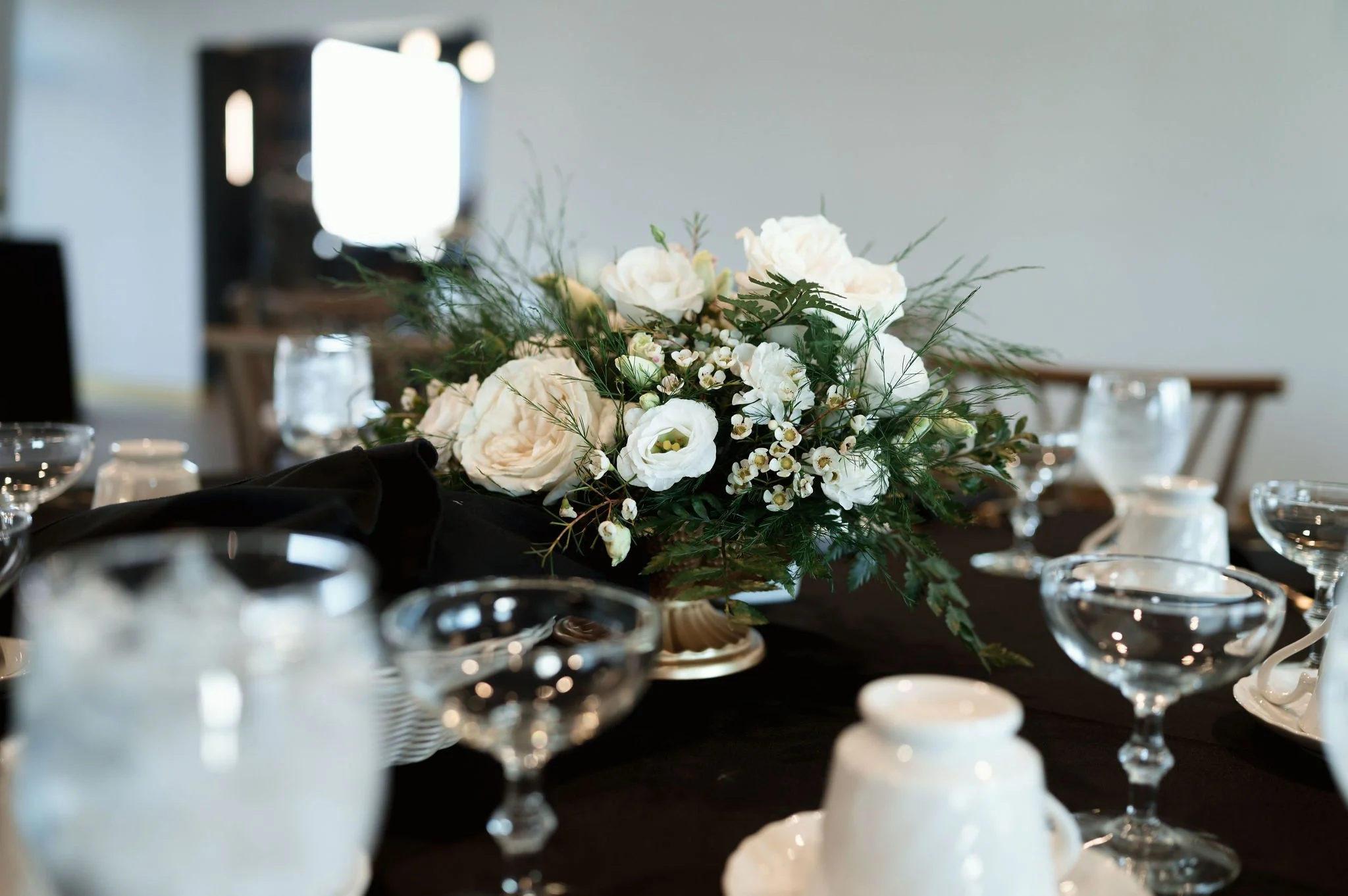A floral centerpiece with white flowers and greenery on a table set with glassware and candles in a dining or event setting.