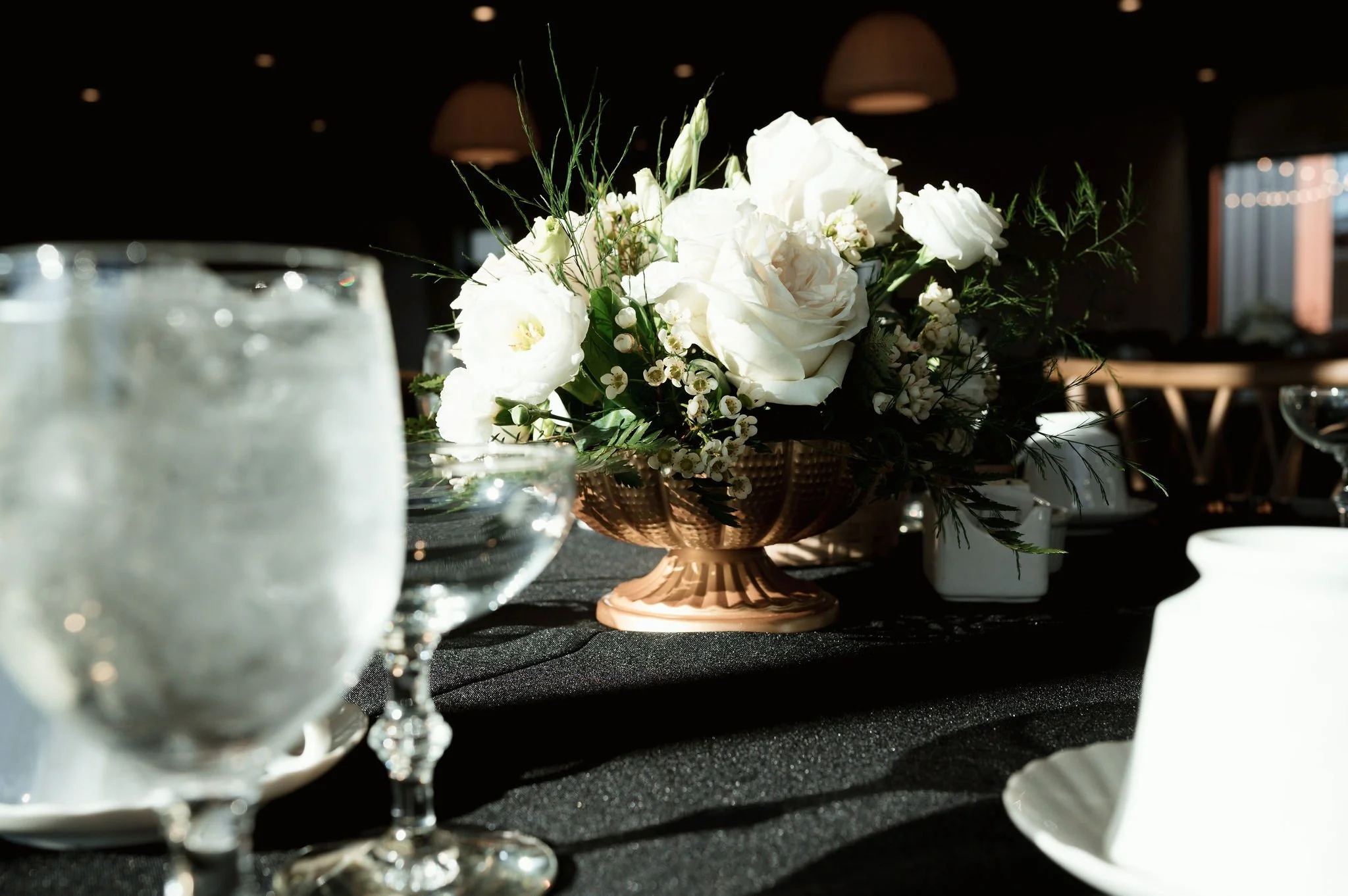 A floral centerpiece with white flowers and greenery in a copper-colored vase on a black tablecloth, with glasses and tableware in the background.