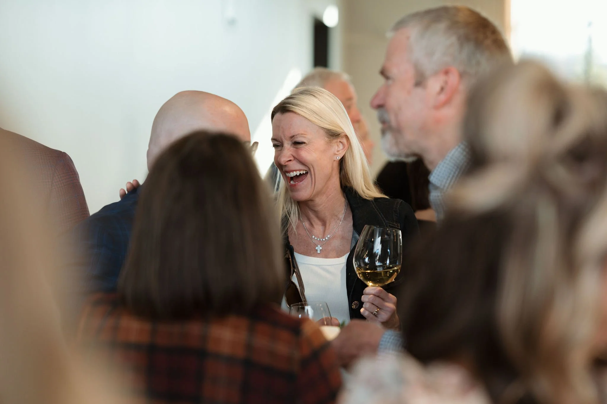 A group of people socializing at a gathering, with a woman holding a glass of white wine, smiling and engaged in conversation.