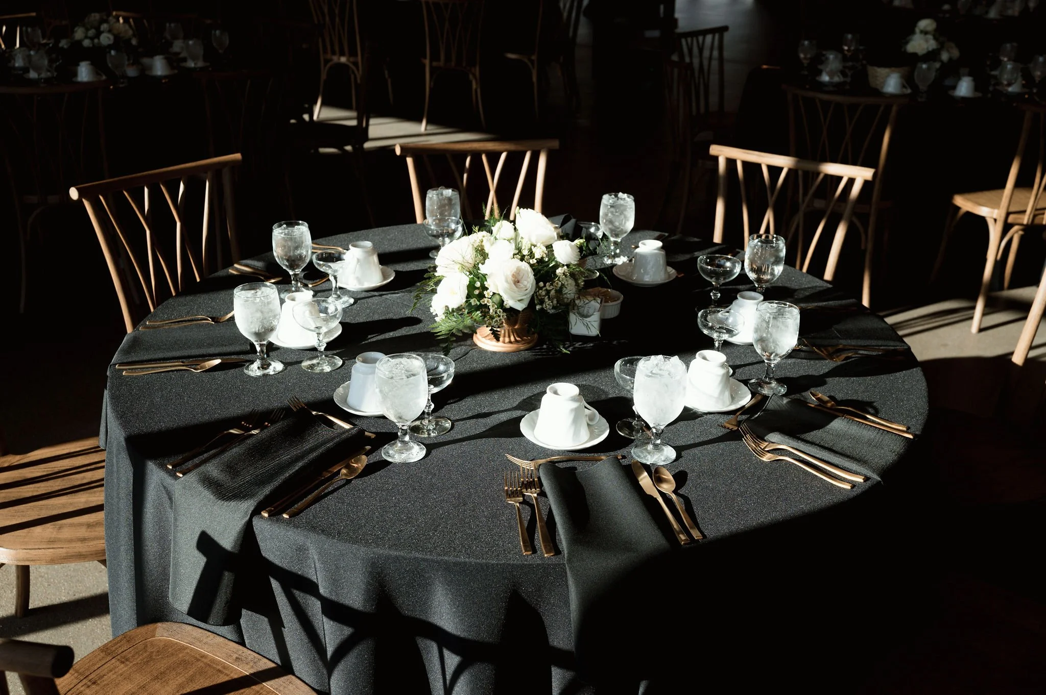 A round table set for a formal event with a black tablecloth, floral centerpiece, water glasses, coffee cups, black napkins, and gold utensils in sunlight, with wooden chairs around.