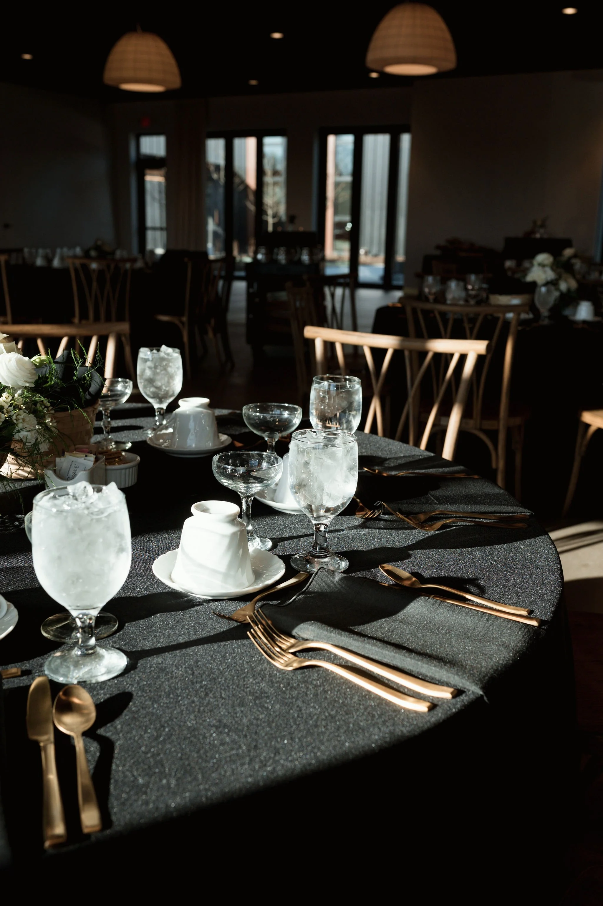 A formal dining setup with black tablecloth, gold utensils, glassware with ice water, white teacups, and a floral centerpiece. Natural light from large windows illuminates the background, which features a modern, elegantly decorated restaurant interi