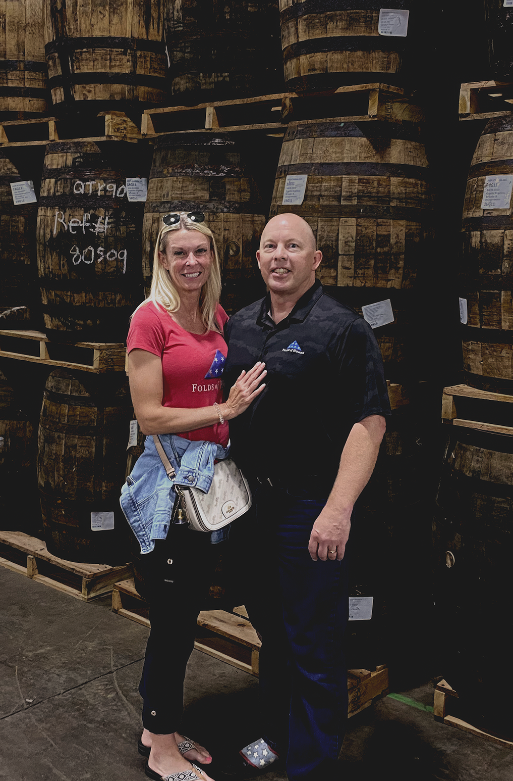 A man and woman standing together in front of large wooden barrels in a warehouse or brewery. The woman is wearing a red T-shirt, black jeans, sandals, and has sunglasses on her head. The man is wearing a black short-sleeve shirt, black pants, and sneakers. They are smiling and posing for the photo.