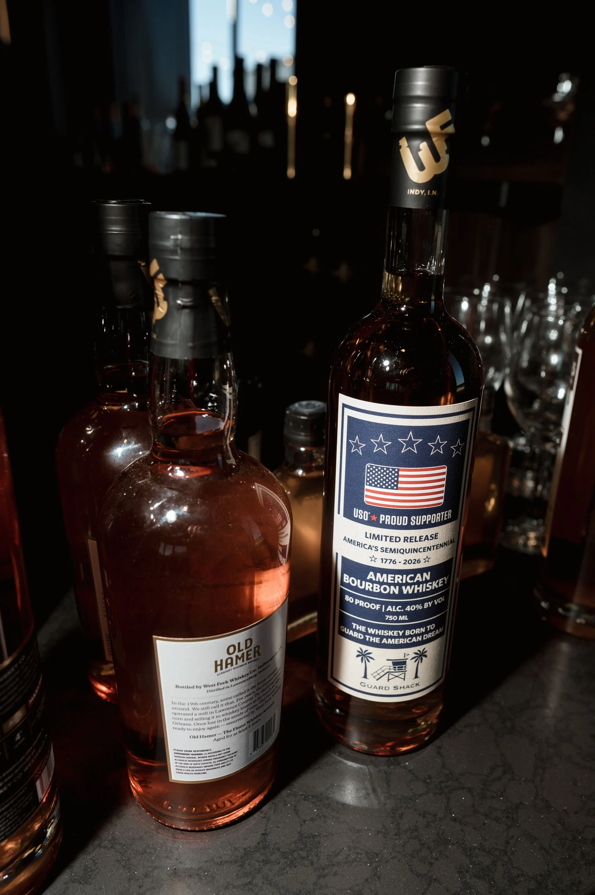 Close-up of two bottles of bourbon whiskey on a dark surface, one labeled 'American Bourbon Whiskey' and the other labeled 'Old Hamer.'