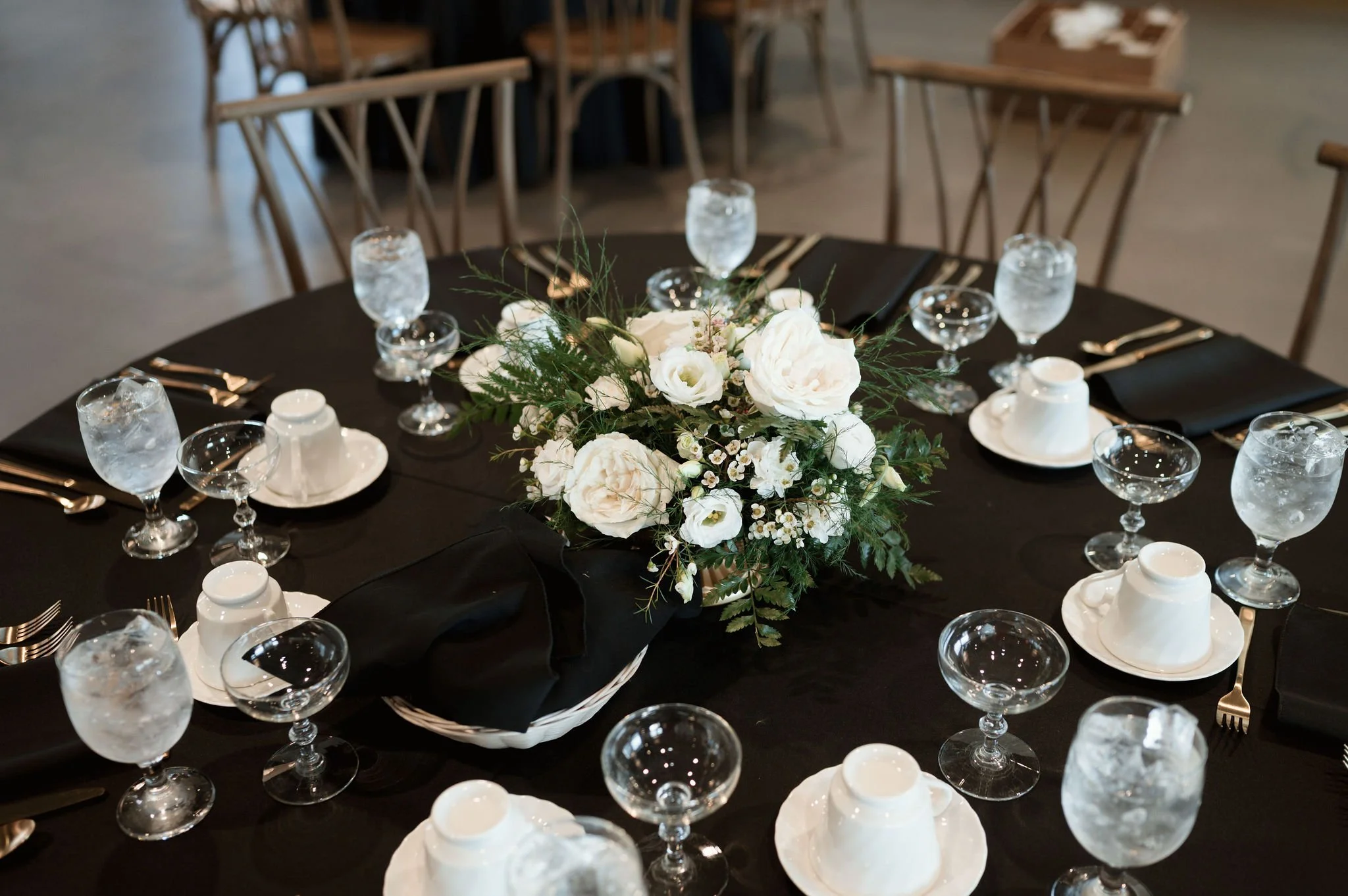 A round table set for a formal event with black tablecloth, floral centerpiece with white flowers and green foliage, multiple glasses of water, white cups and saucers, black napkins, and silverware, in a venue with wooden chairs.