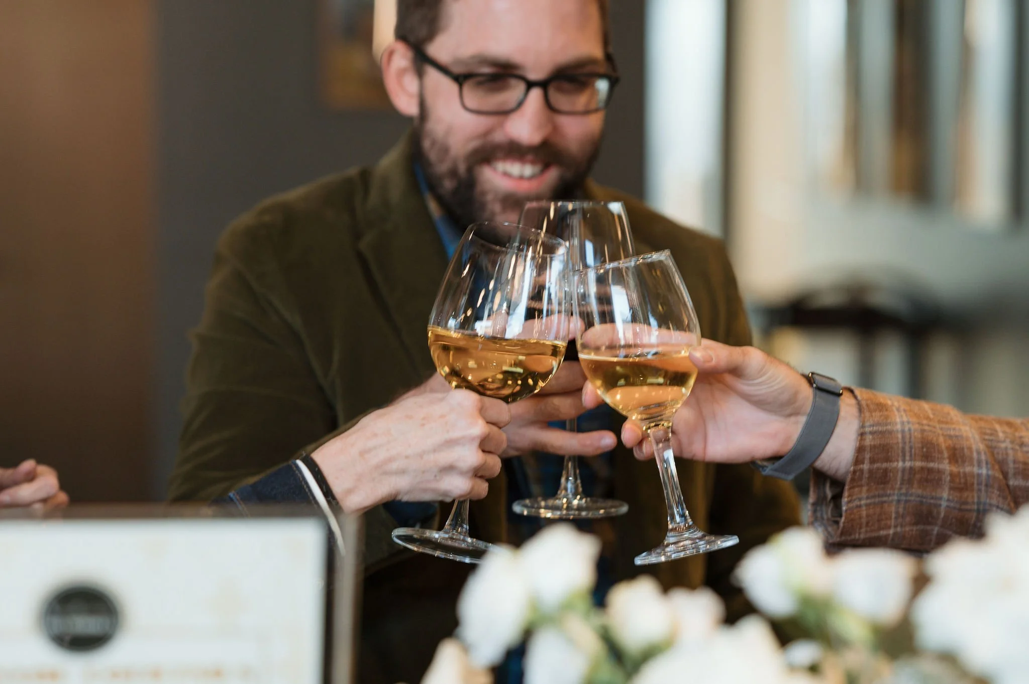 A man with glasses and a beard smiling as he toasts with three glasses of white wine, surrounded by others in a social gathering.