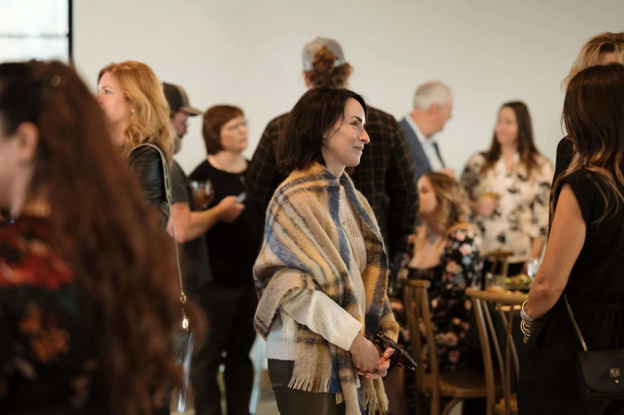 A woman with dark hair wearing a beige, blue, and brown striped scarf and beige sweater, smiling and holding sunglasses, standing among a group of people indoors.