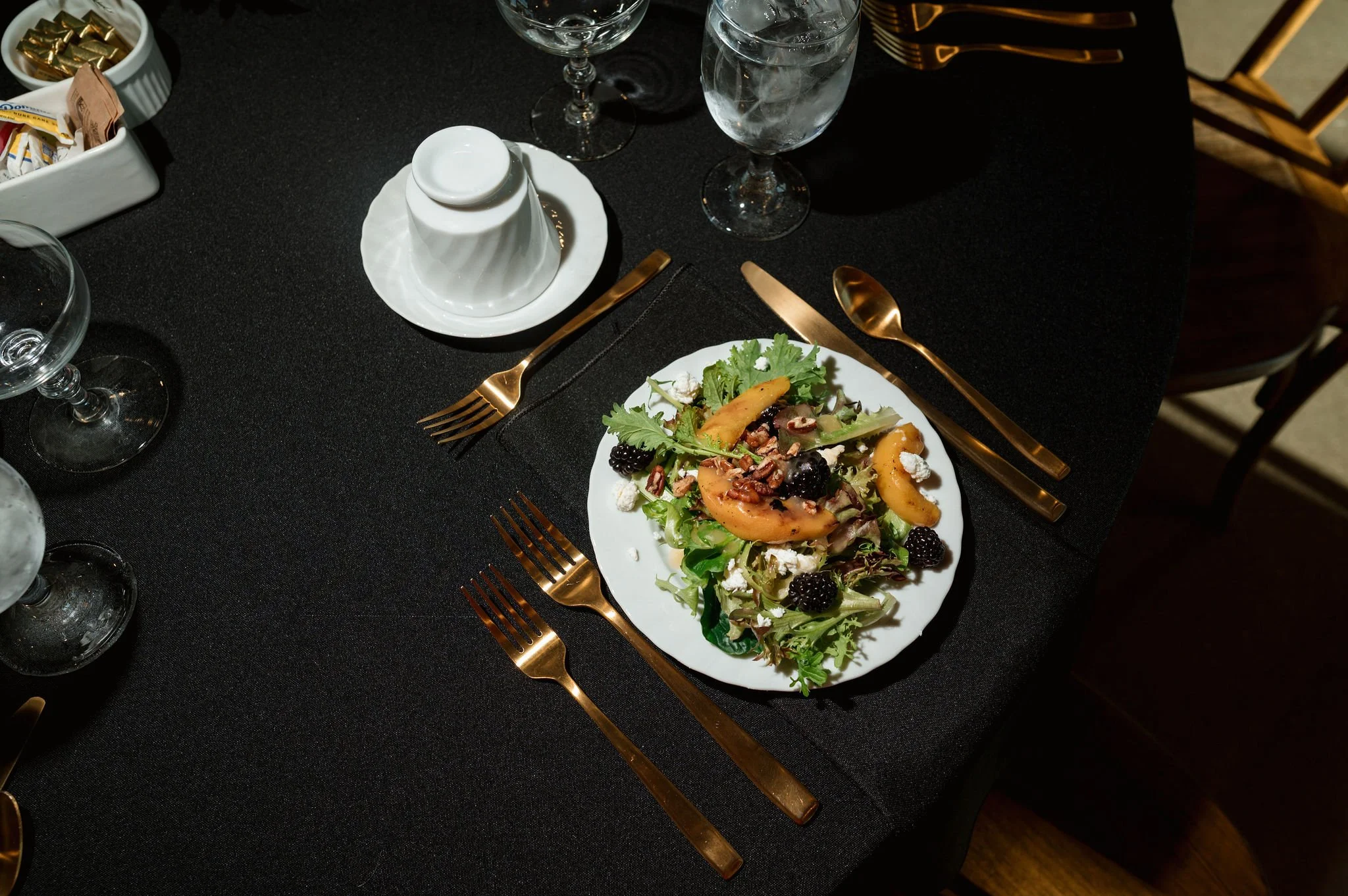 A table set for a meal with a plate of salad, drinking glasses, and gold-colored utensils on a black tablecloth.