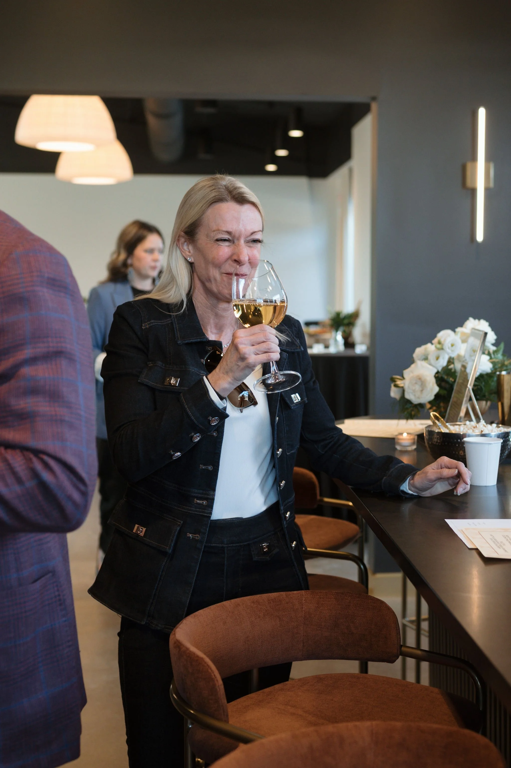 Woman with blonde hair smiling and holding a glass of white wine in a modern cafe or restaurant setting.