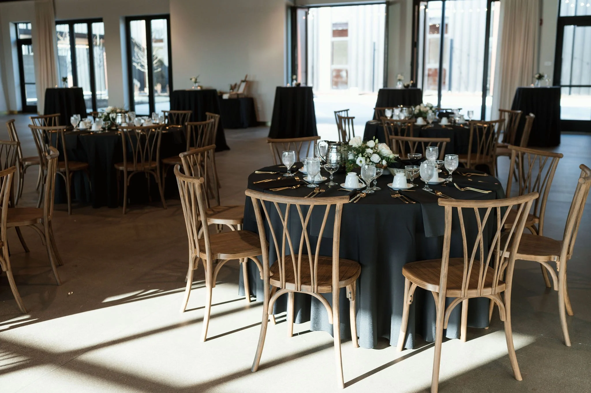 A banquet hall set up with round tables covered in black tablecloths, decorated with white floral centerpieces, glassware, and gold cutlery, with natural light coming through large windows.
