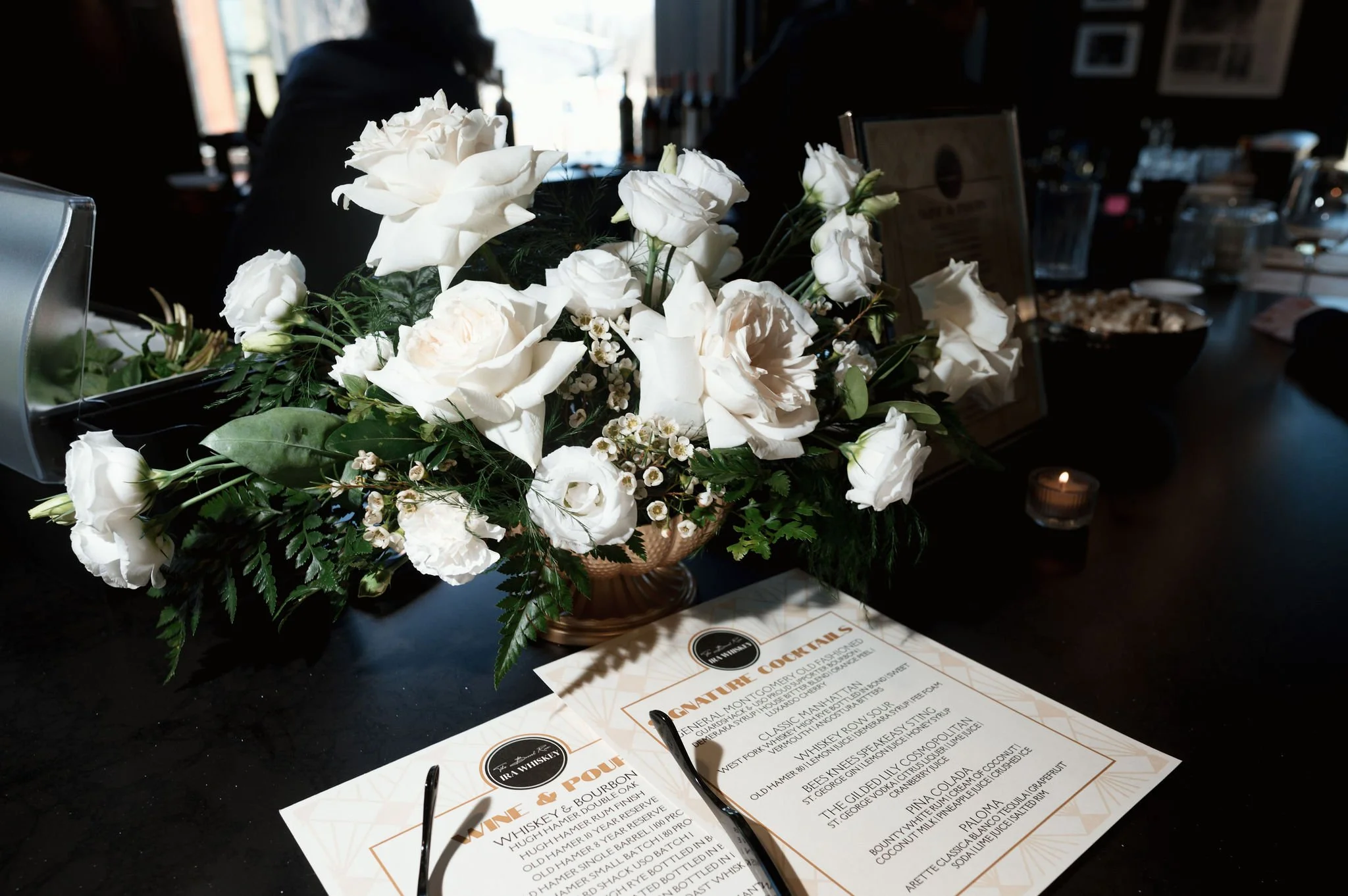 A bouquet of white flowers, including roses and lilies, placed on a restaurant or bar counter. In the background, there is a blurred person and a menu or drink list on the table.