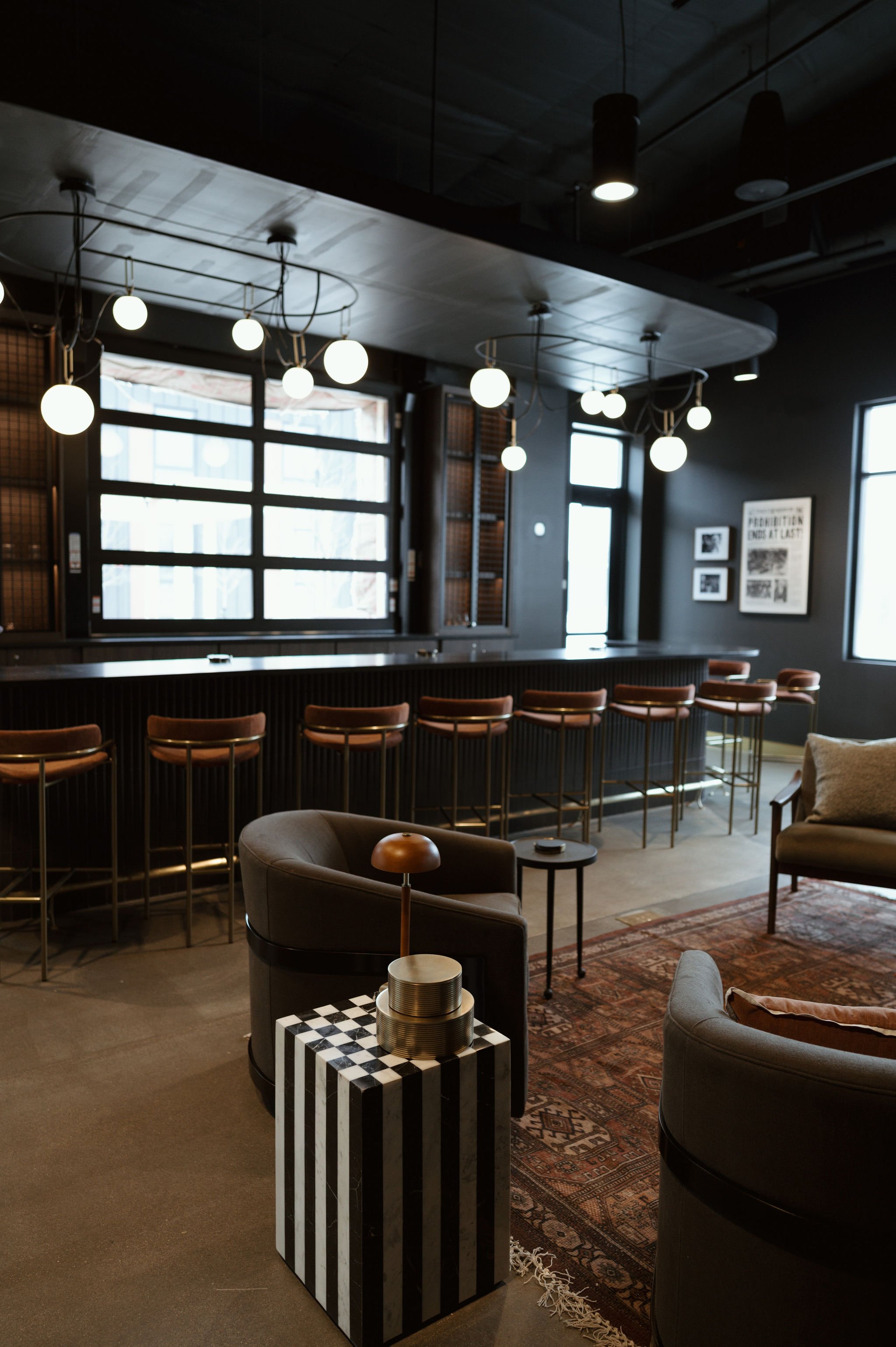 A modern lounge area with a bar counter, black walls, and black ceiling. Upward hanging spherical white lights from black cords. Two armchairs and a sofa around a coffee table with decorative items. Large window with grid panes and framed pictures on
