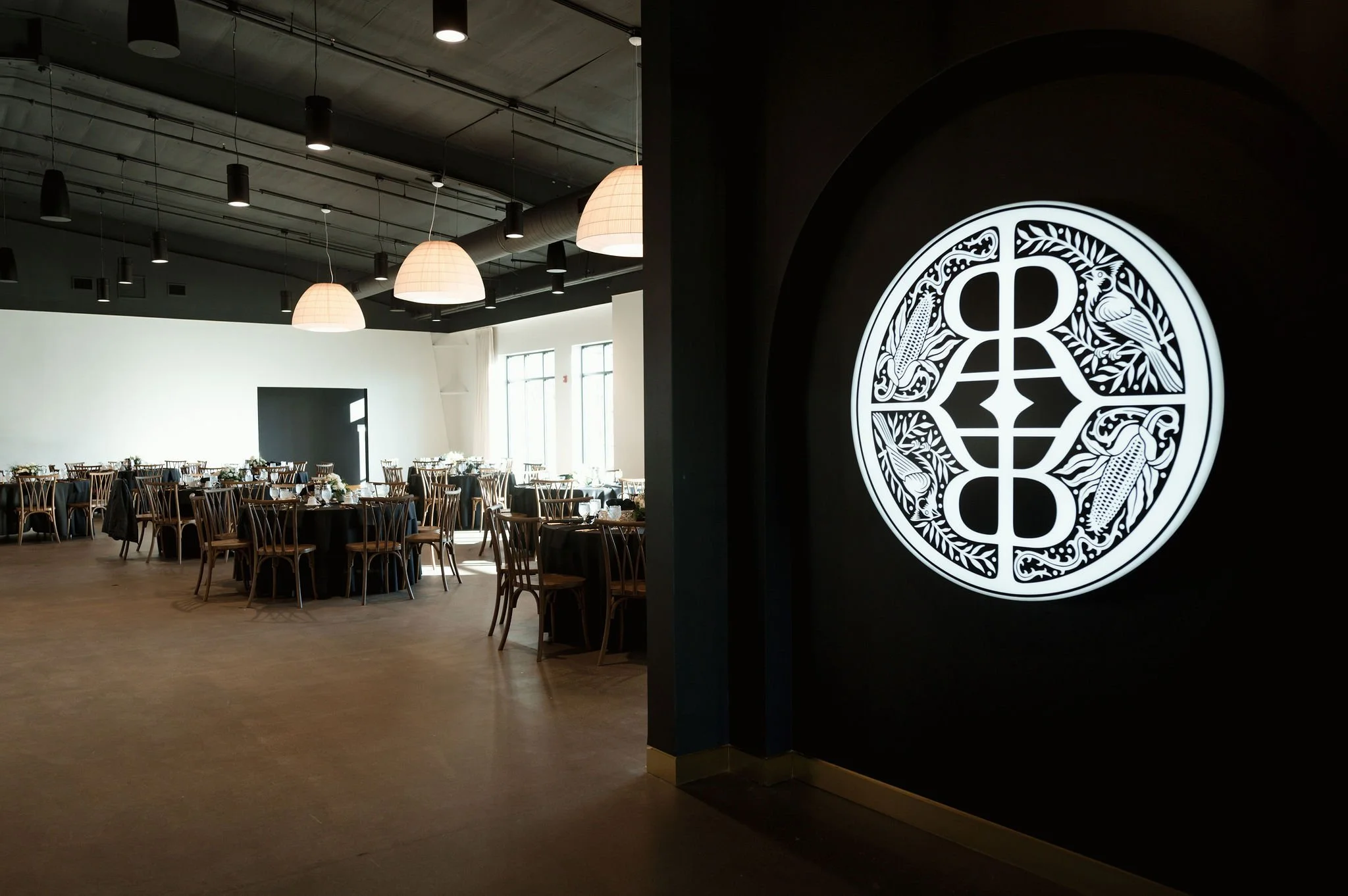Interior of a modern event space or restaurant with round tables covered in black tablecloths, wooden chairs, and large window letting in natural light. There are hanging light fixtures on the ceiling and a large illuminated circular logo on a dark w