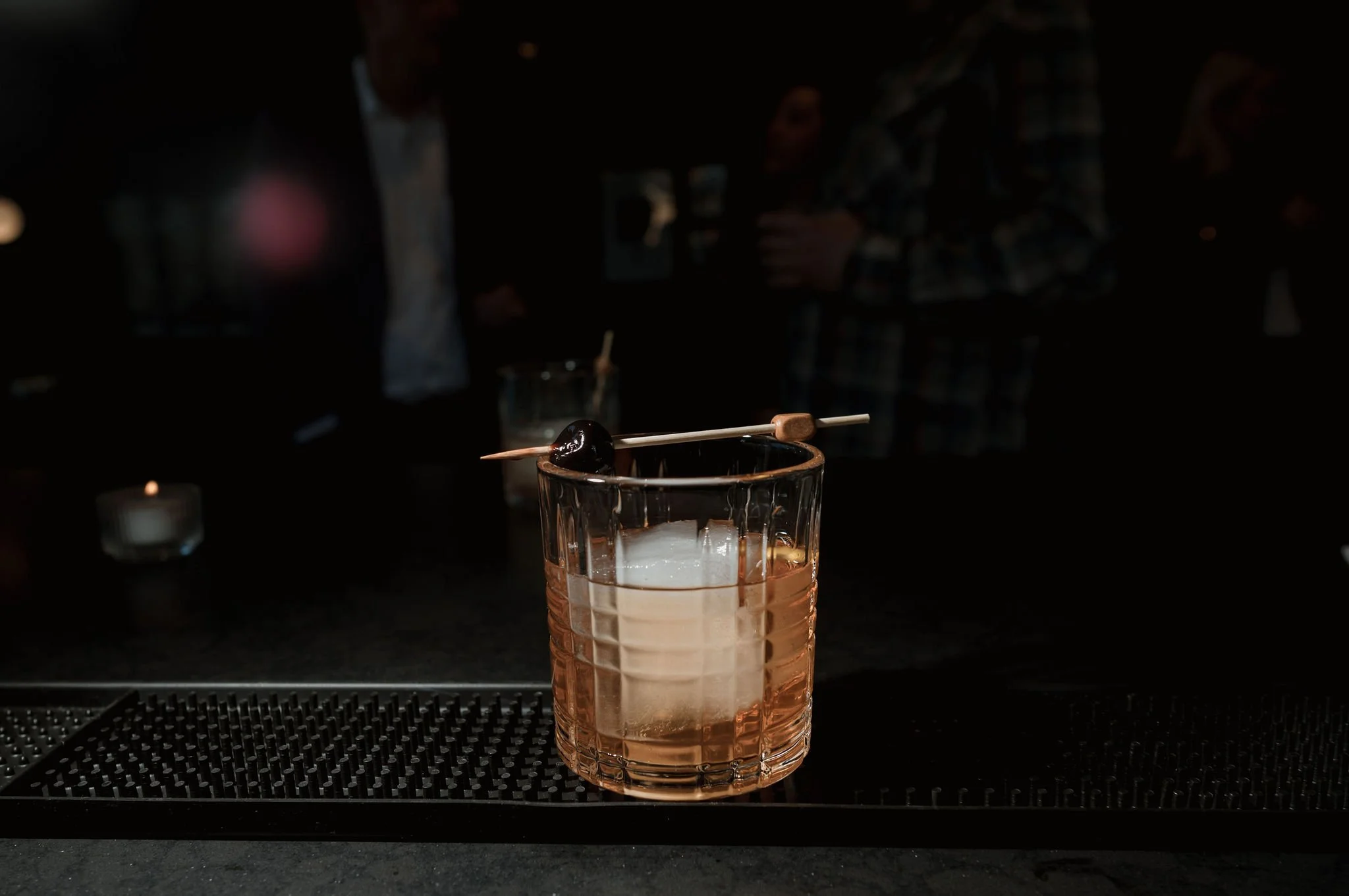 A glass with a pinkish-orange drink and an ice cube, garnished with a cherry on a cocktail pick, sits on a bar counter in a dimly lit setting.