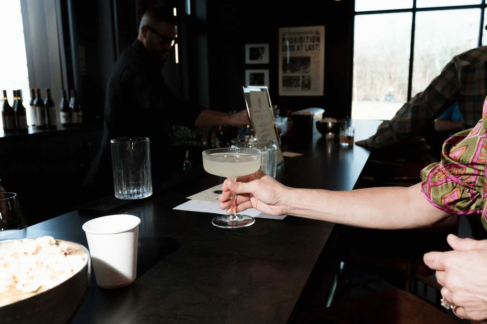 Person holding a cocktail glass with a light-colored drink inside at a bar or social gathering.