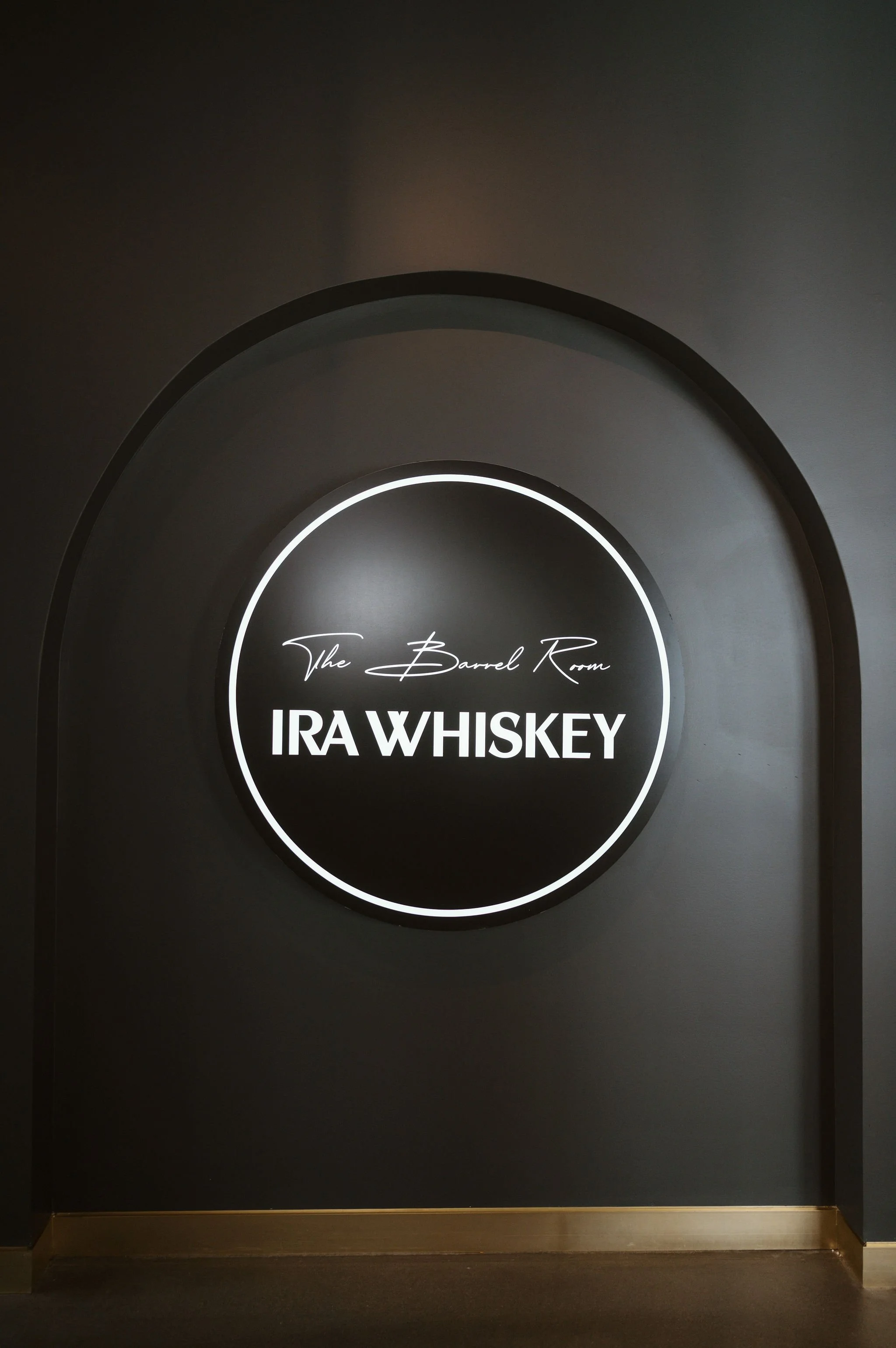A black circular sign with white neon lighting that reads "The Barrel Room IRAX WHISKEY" on a gray wall.