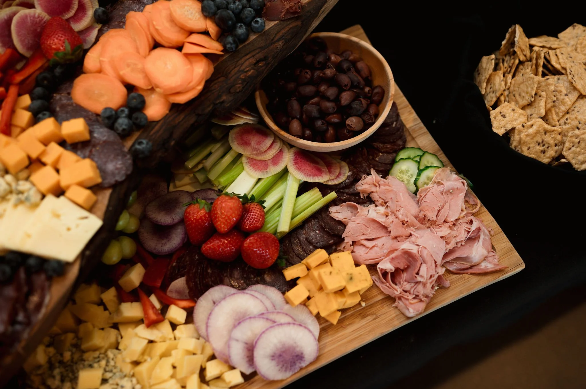 Assorted cheeses, fresh strawberries, radishes, cucumber slices, sliced meats, black grapes, blueberries, granola, and crackers arranged on a platter.