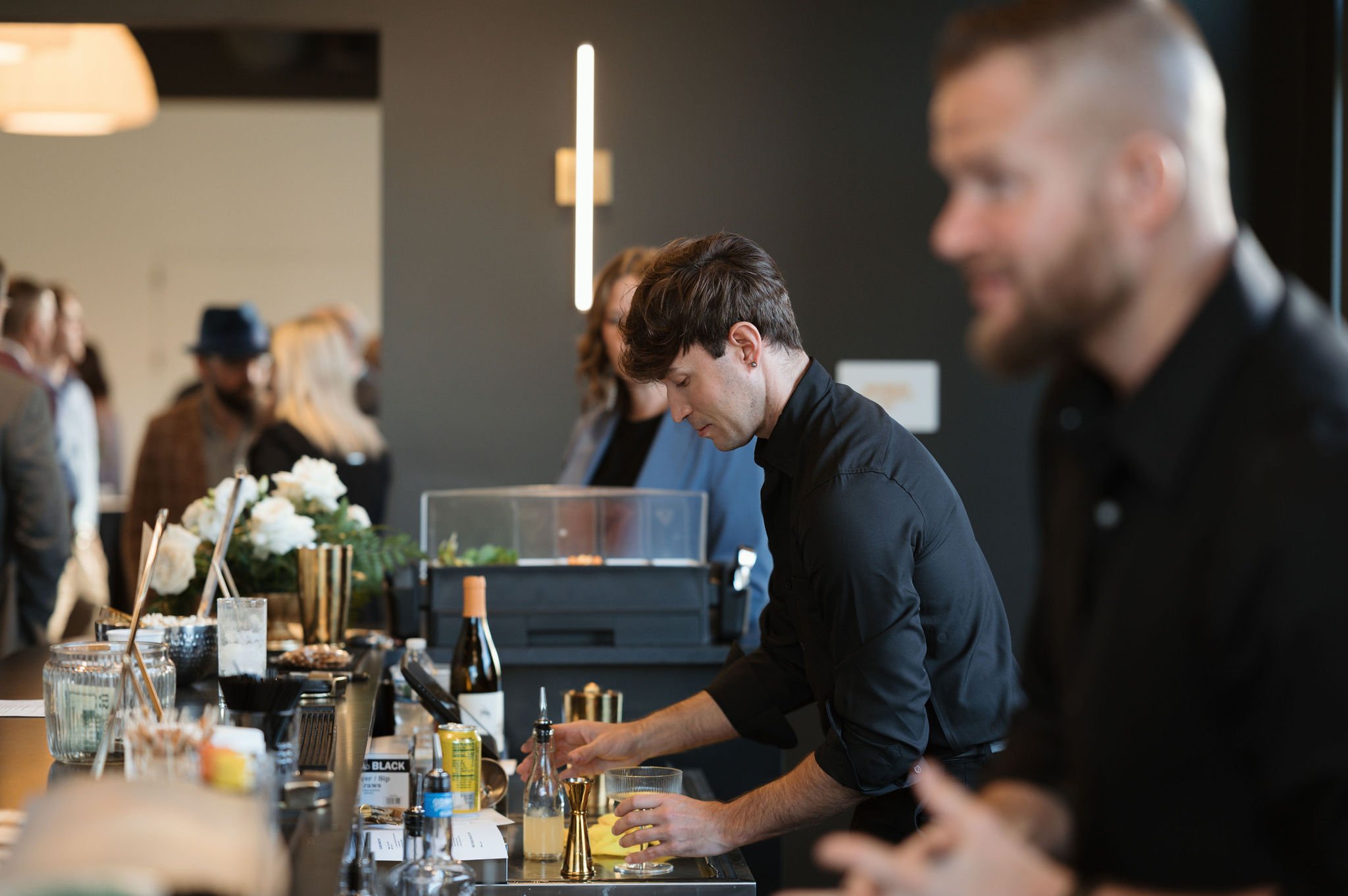 Bartender preparing a drink at a bar counter with various bottles and glasses, surrounded by patrons in a modern, stylish bar or restaurant.