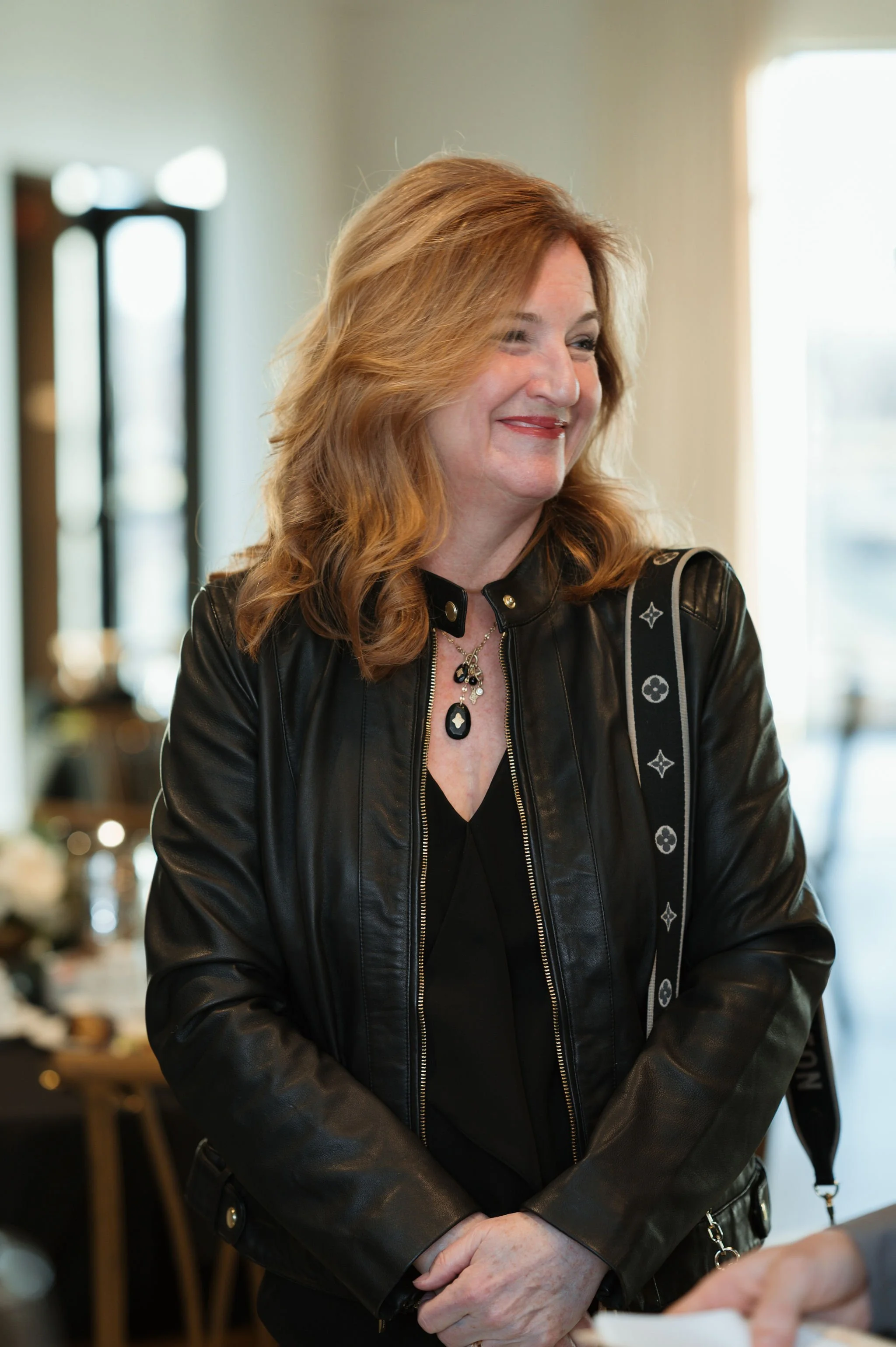 A woman with red hair wearing a black leather jacket and jewelry, smiling indoors.