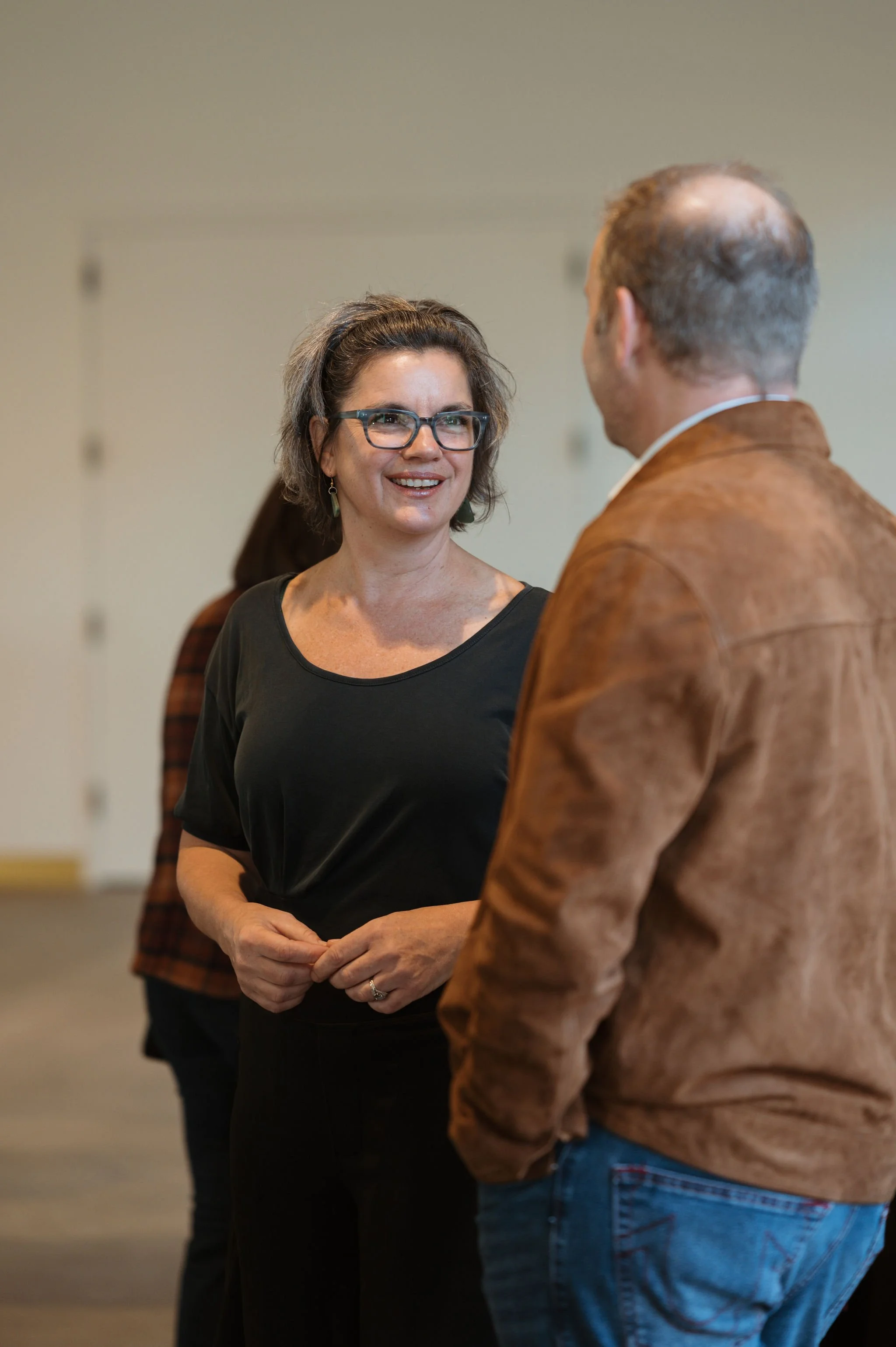 A woman with glasses and a black top smiling and talking to a man wearing a brown jacket and jeans at an indoor event, with another person in the background.