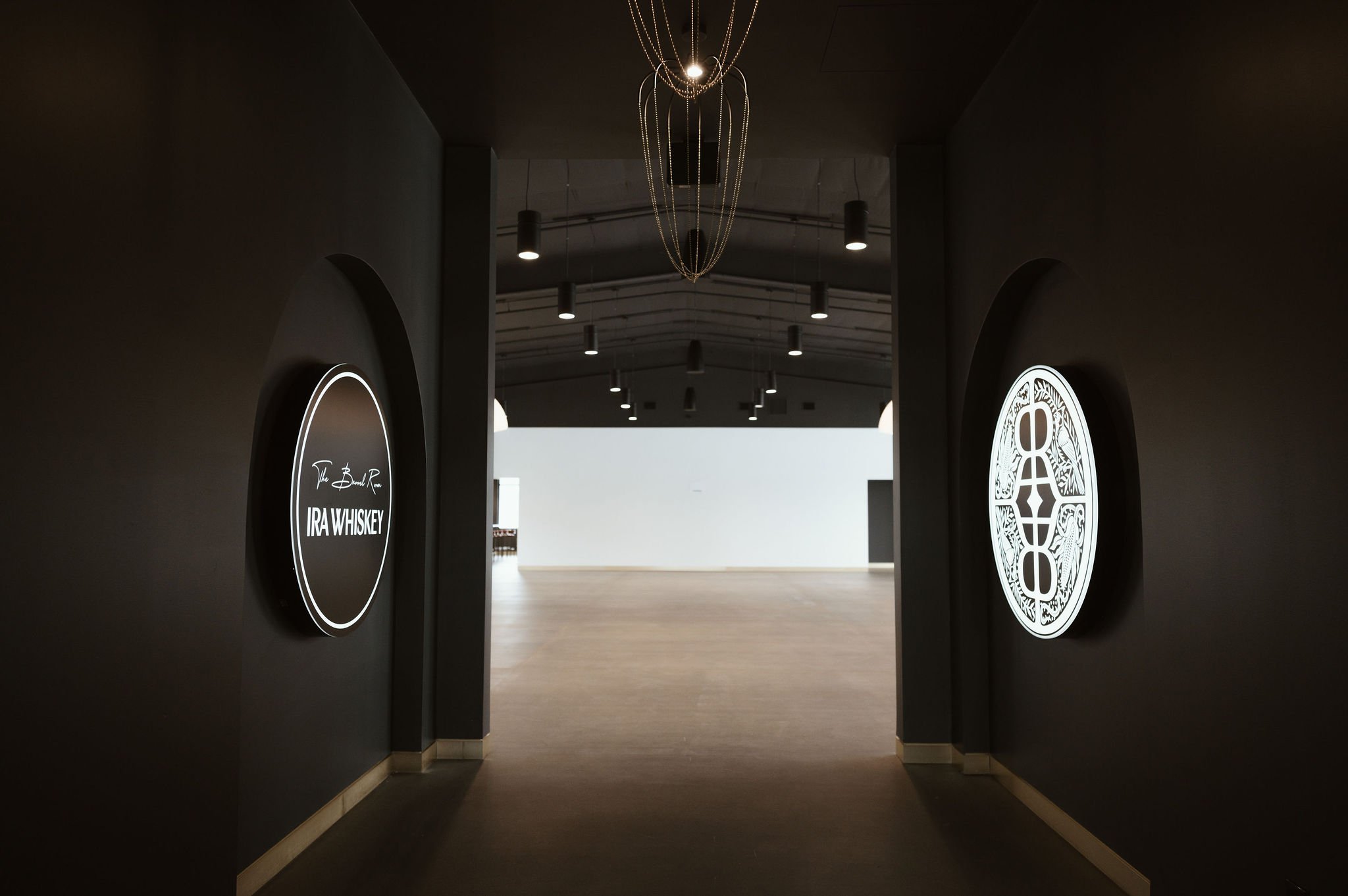 An indoor hallway with dark walls featuring illuminated signs for IRA Whiskey. The hallway is dimly lit with ceiling lights and a decorative chandelier at the center ceiling. The corridor opens to a brightly lit area in the background.