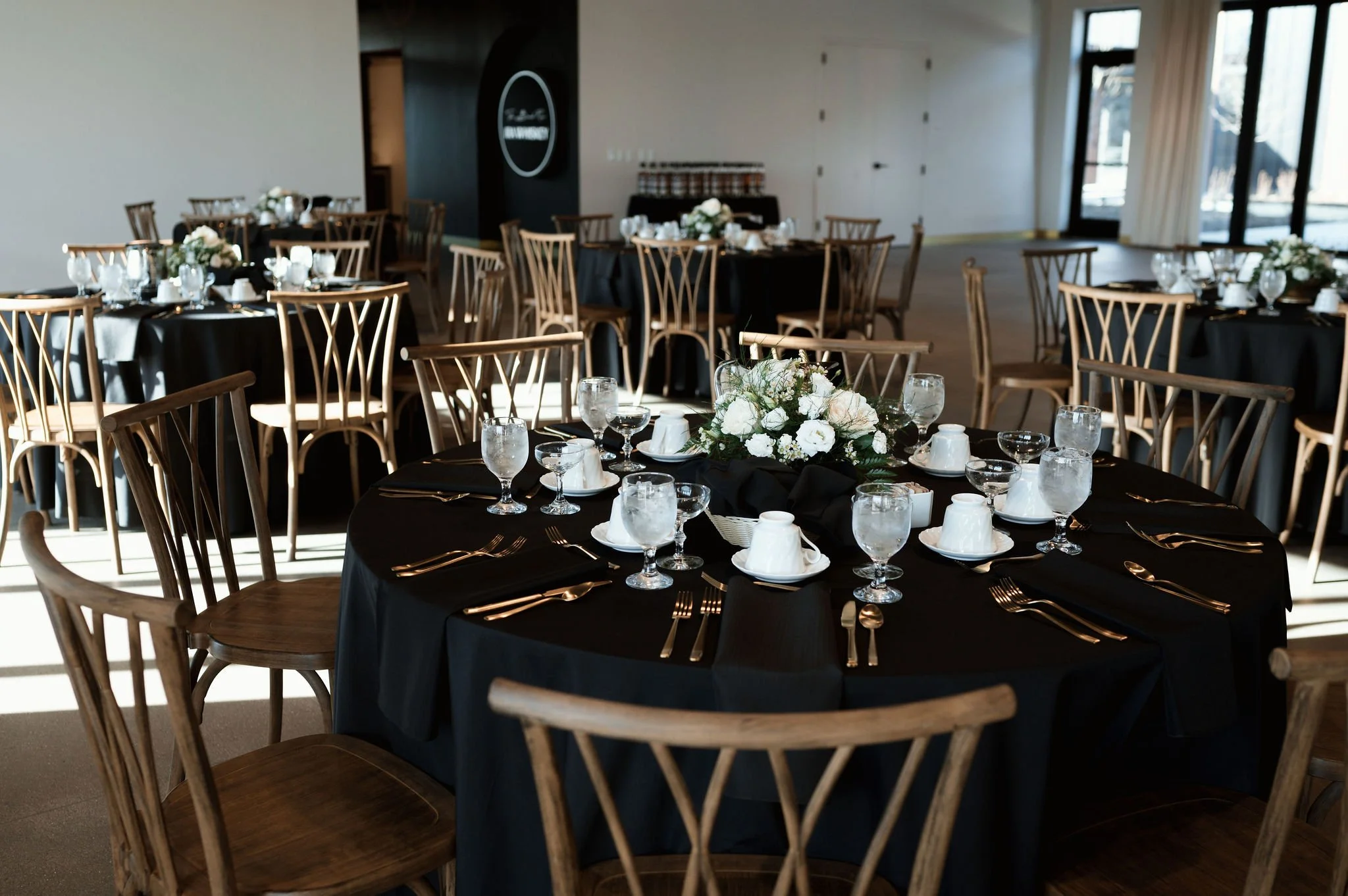 Round banquet tables with black tablecloths and elegant place settings, including glassware, white cups, and wrapped utensils, decorated with flower centerpieces, in a well-lit room with large windows and a neutral color scheme.