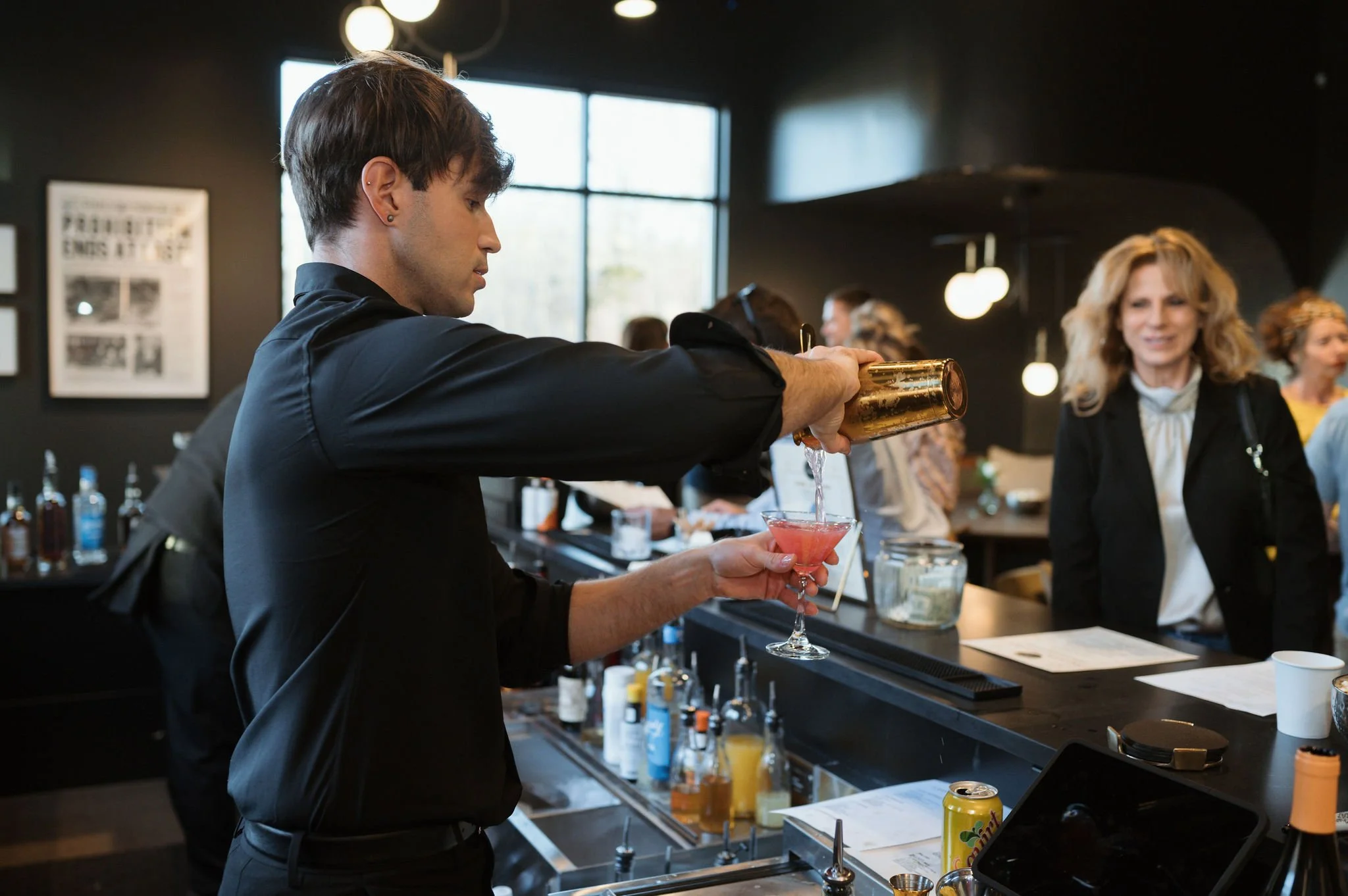A bartender pouring a pink cocktail into a glass at a bar with several customers in the background.