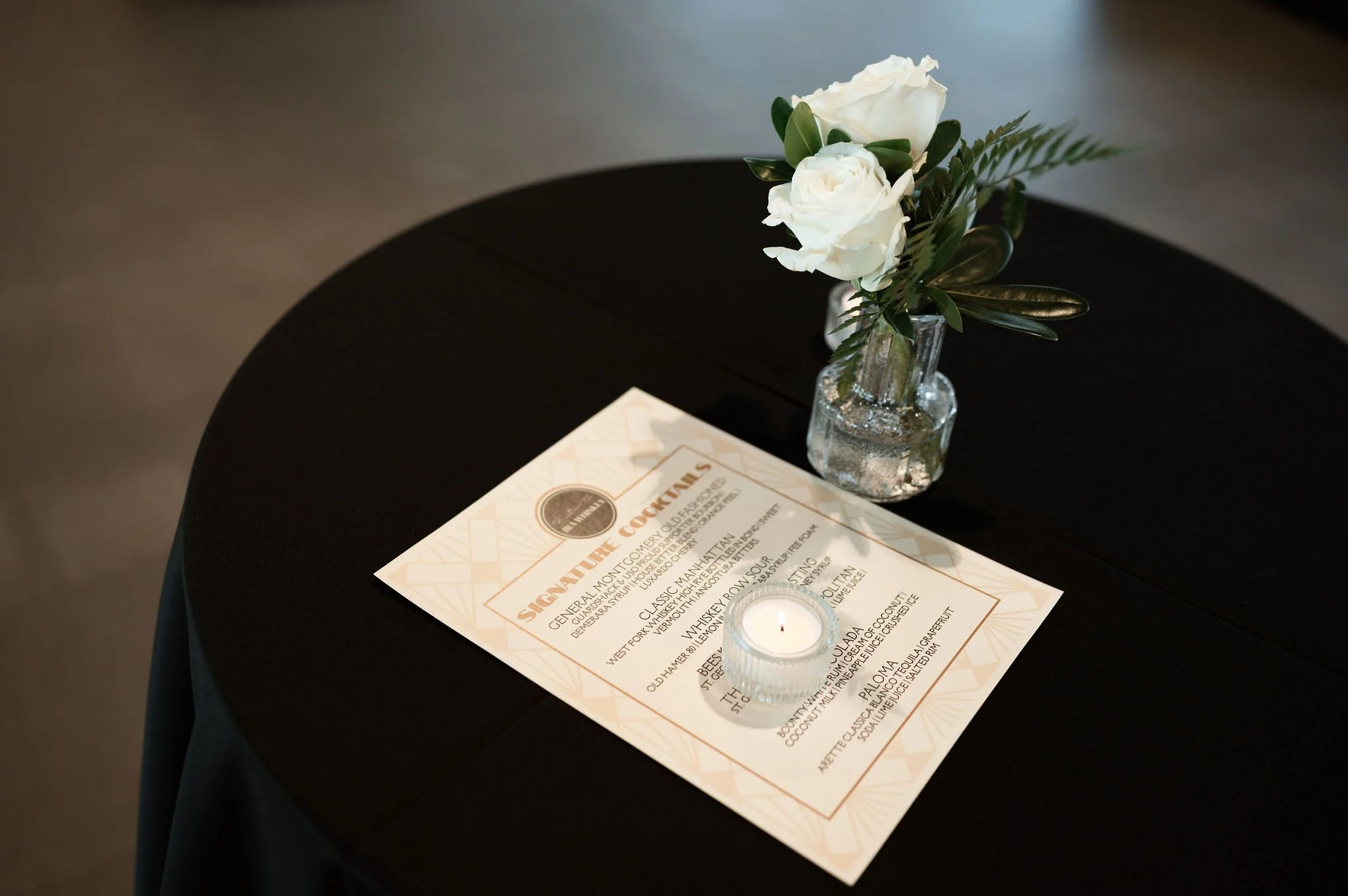A black table with a glass vase containing white roses and green leaves, a candle, and a printed menu or program.