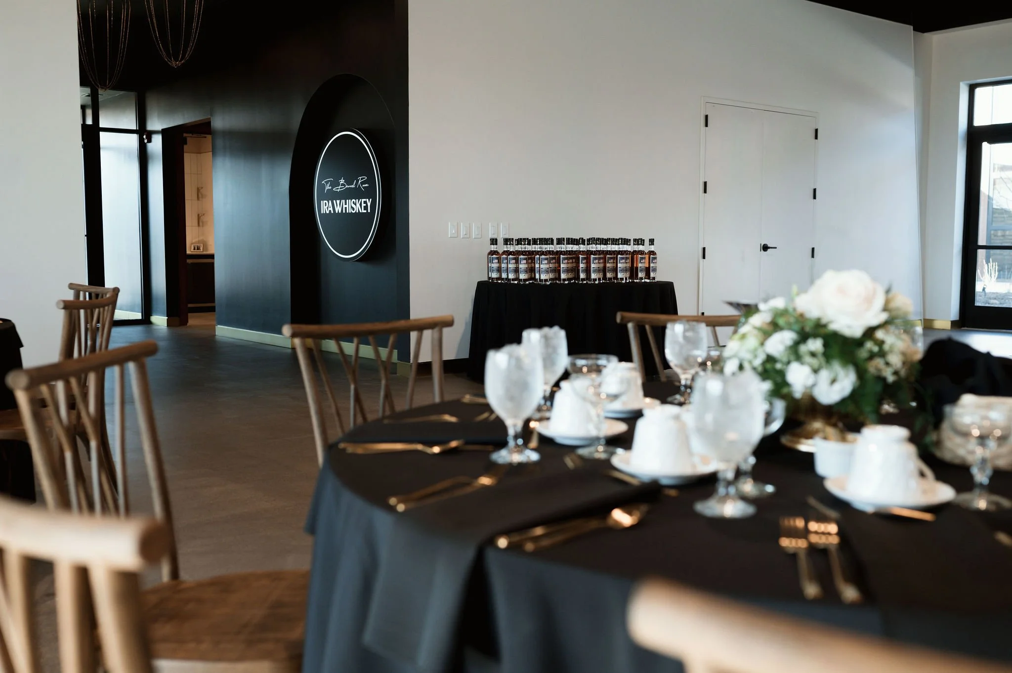 Interior of a modern event space with tables set for a meal, including plates, glasses, and a floral centerpiece, with a black wall featuring a round illuminated sign that reads 'IRA WHISKEY' and bottles placed on a black table.