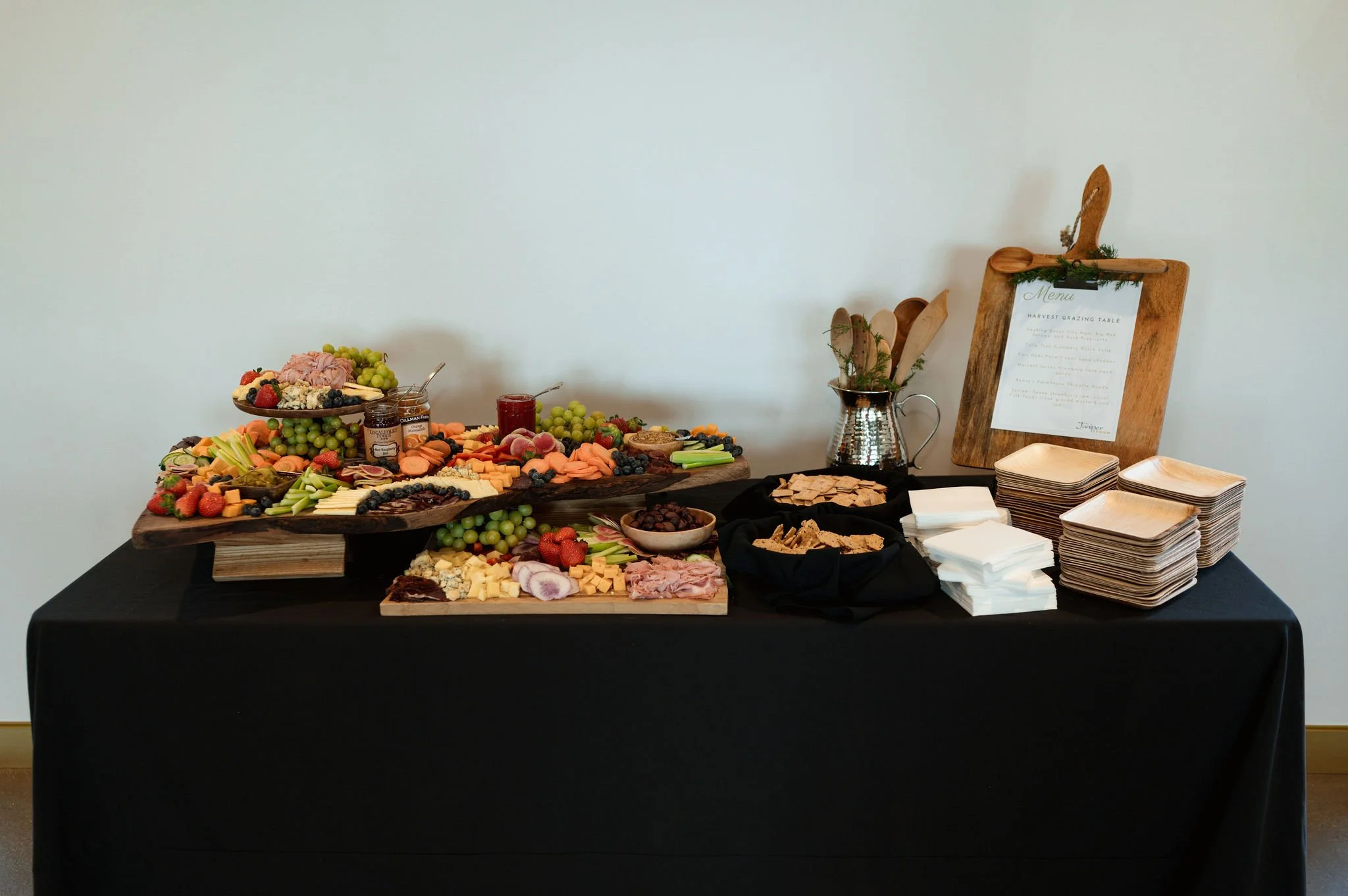 A table with a black tablecloth holding a cheese and fruit platter, jars of jam, and various snacks; a smaller cheese board and plates of crackers and napkins are also on the table; a menu sign is in the background.