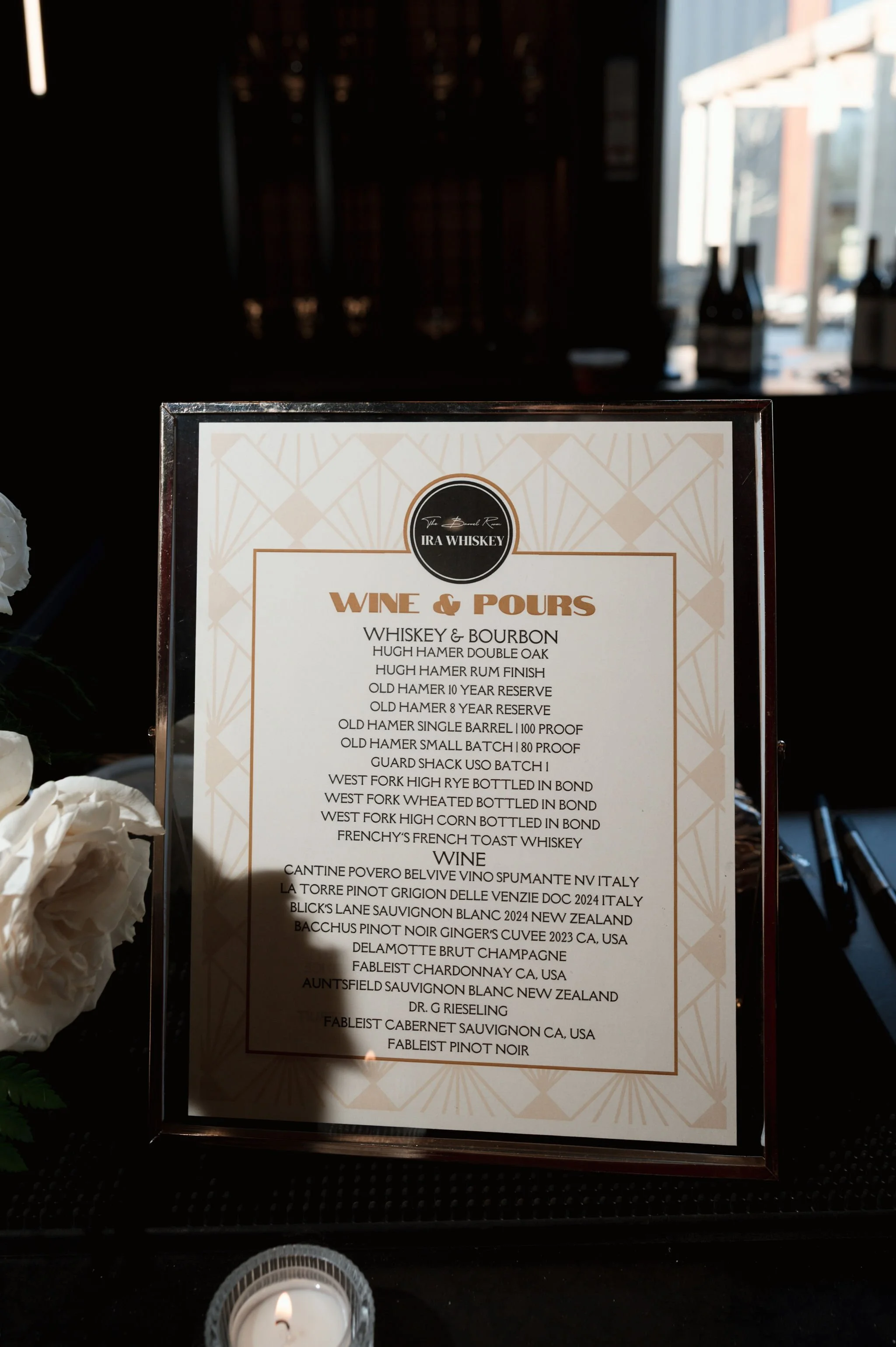 A menu titled 'Wine & Pours' featuring whiskey, bourbon, and wine options, displayed inside a frame on a table, with a lit candle in a small glass holder in front of it.