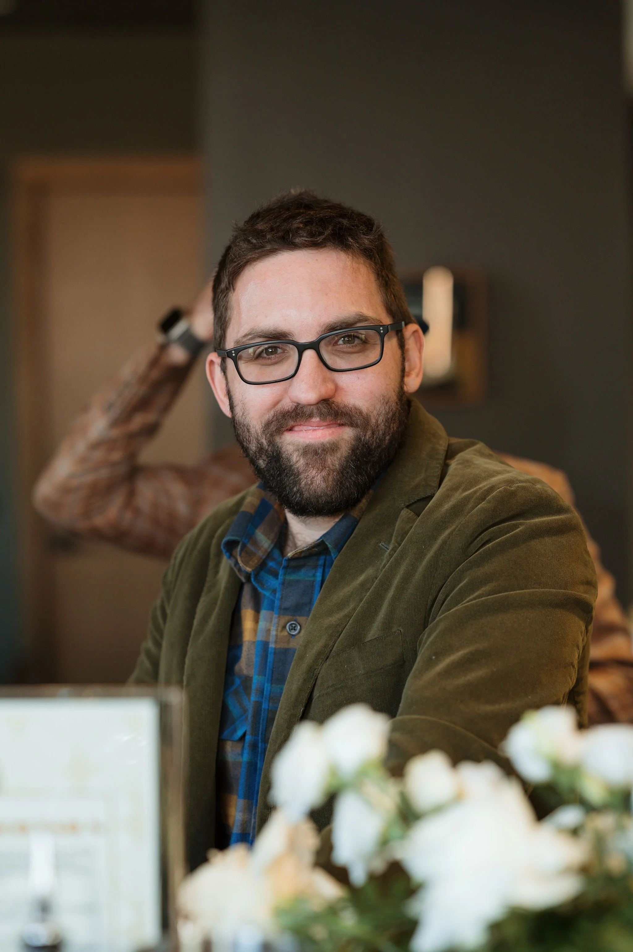 A man with glasses and a beard smiling at the camera, sitting in an indoor setting, with another person in the background and white flowers in the foreground.
