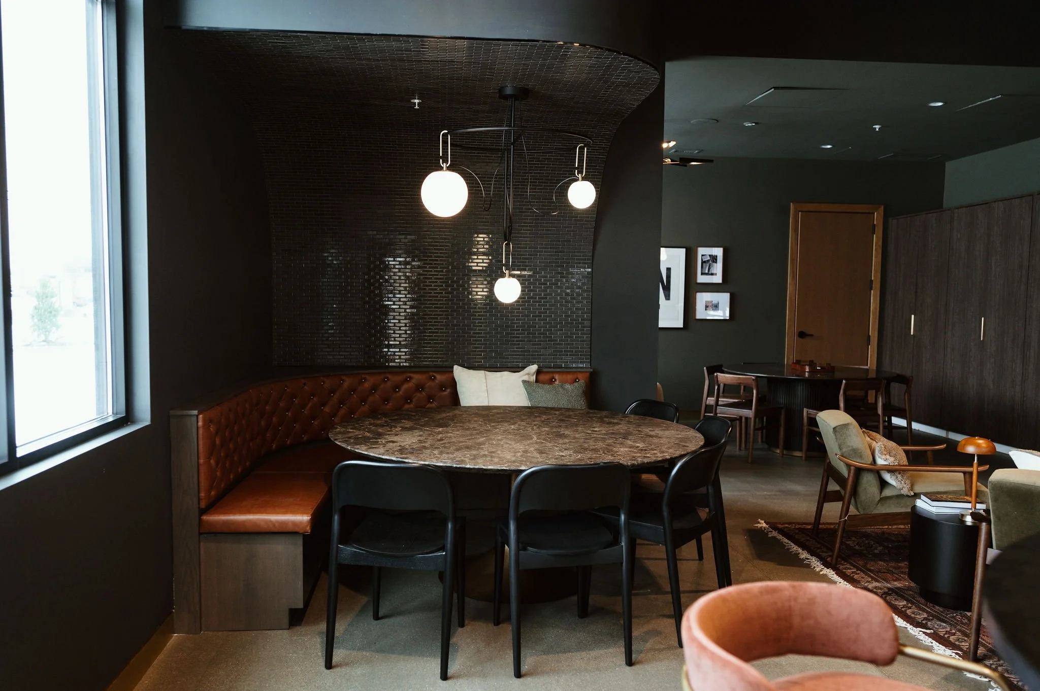 Interior of a modern lounge or cafe with dark walls and wooden furniture, featuring a large round marble table, black chairs, a velvet upholstered booth seat, and various framed pictures on the wall. Pendant lights hang from the ceiling near a window
