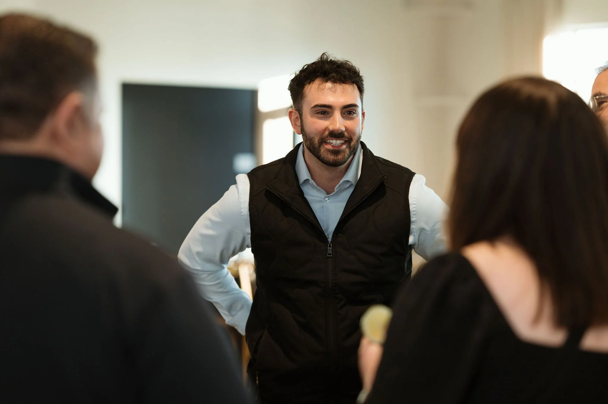 A man with dark, curly hair and a beard smiling and talking to a group of people in a bright room.