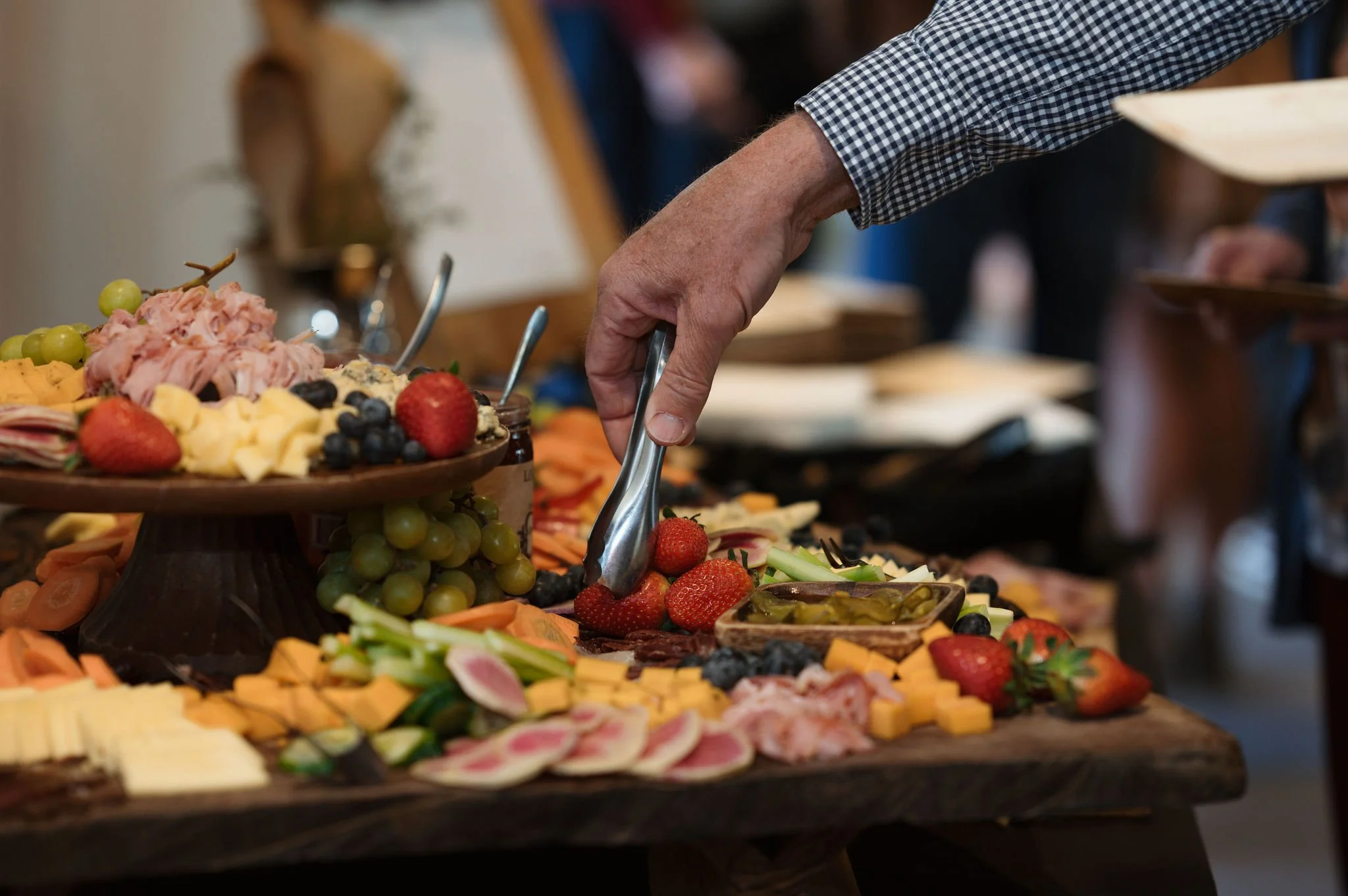 Close-up of a person's arm reaching for strawberries at a buffet table filled with various types of fruit, cheese, and meats.
