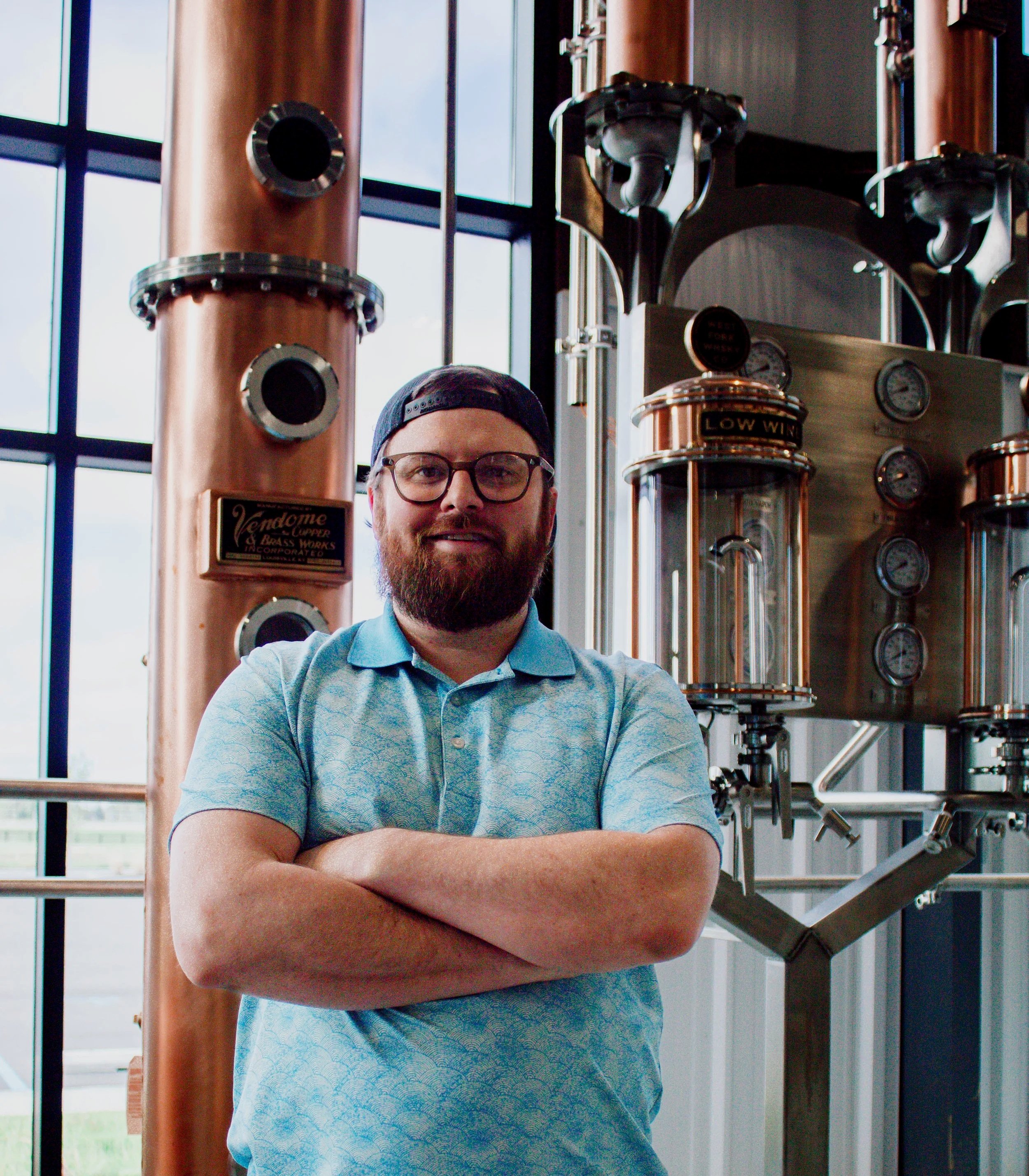 A man with glasses and a beard, wearing a blue patterned polo shirt and a baseball cap, standing in front of a large window and a copper and black industrial machine with gauges and pipes.